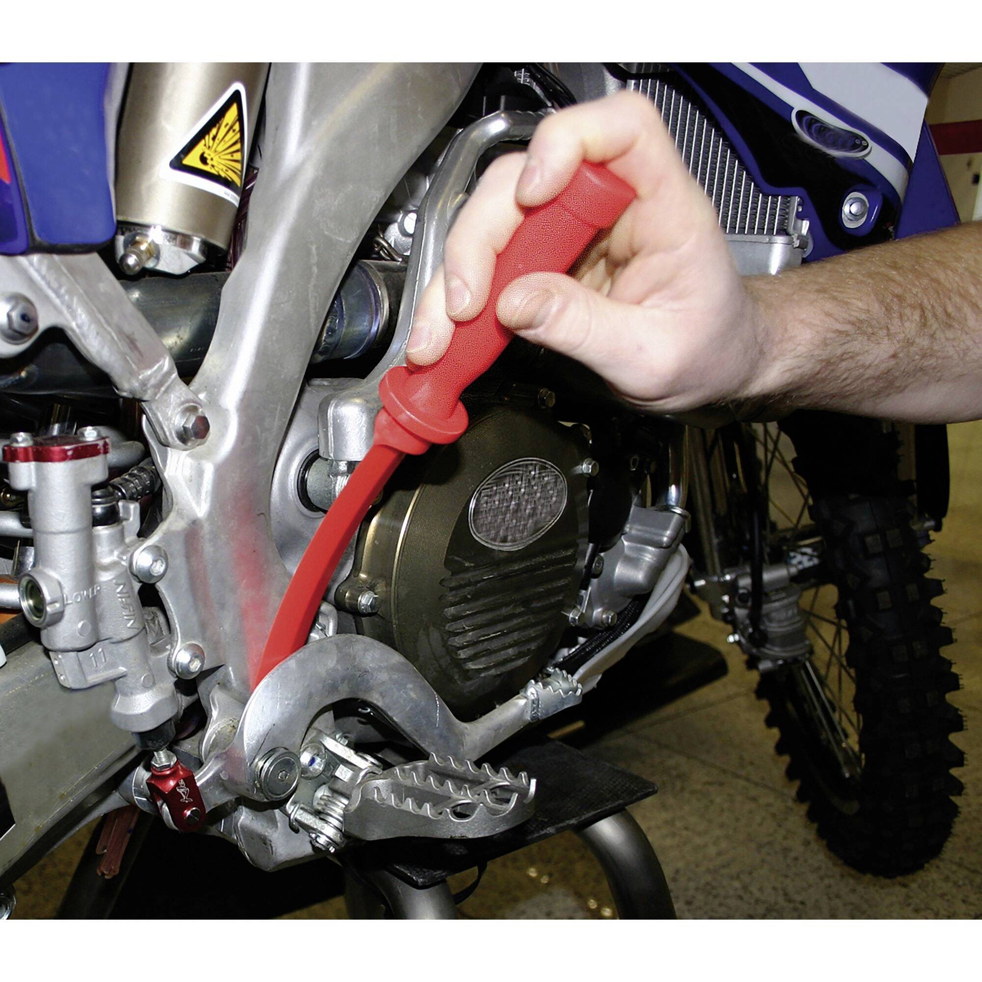 Een persoon houdt een rood gereedschap vast en werkt aan een motorfiets. De focus ligt op de voetpedaal en de kettingslot van de motorfiets.