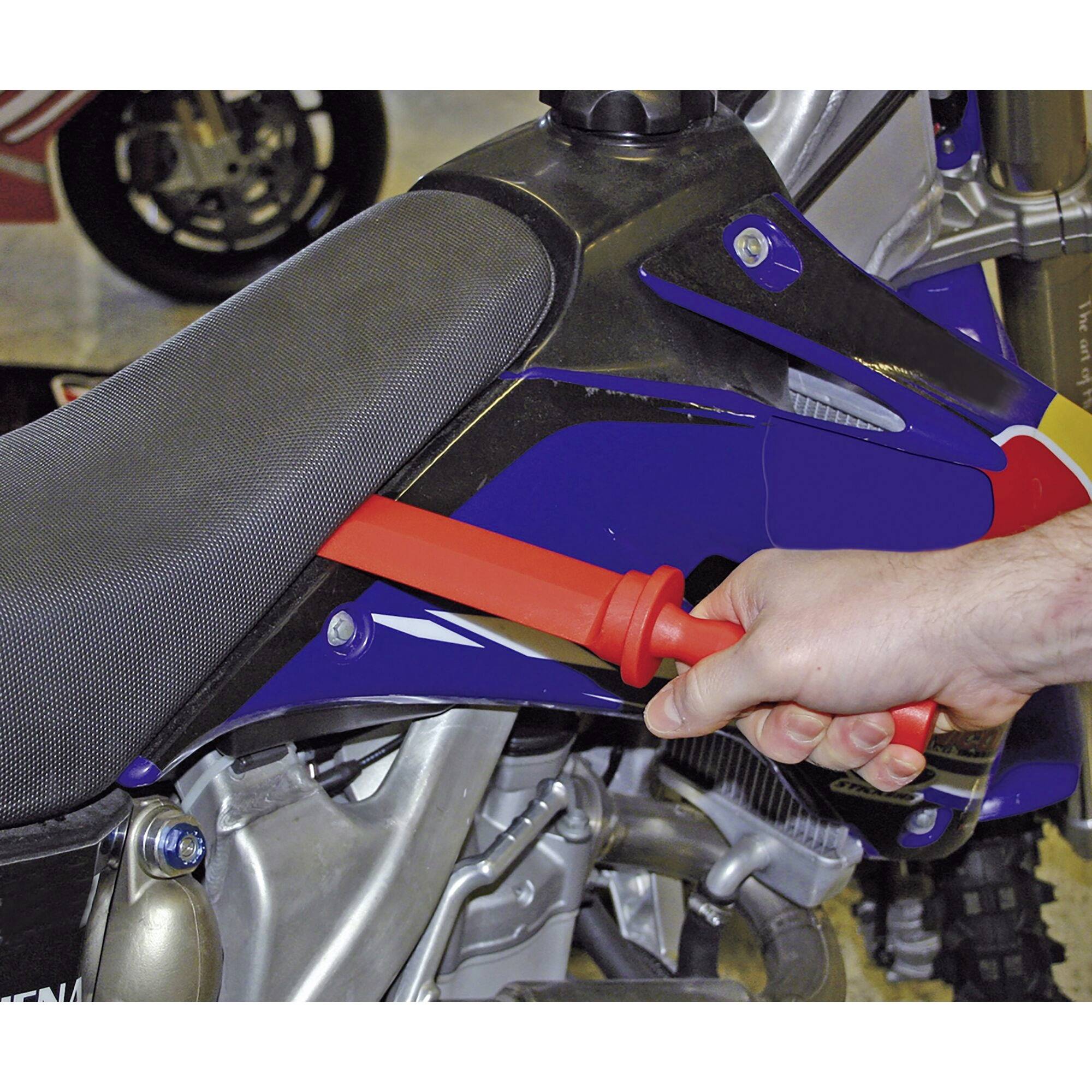 Een persoon controleert de bandenspanning van een motorband met een rood meetapparaat. De motorfiets heeft een blauwe kuip.