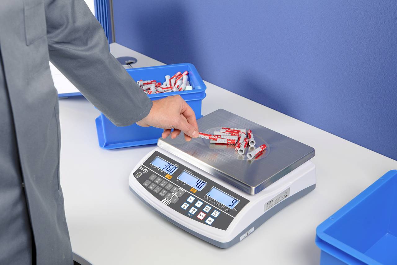 Een persoon weegt metalen onderdelen op een digitale weegschaal; de display toont 360 gram. De weegschaal staat op een witte tafel naast blauwe containers.