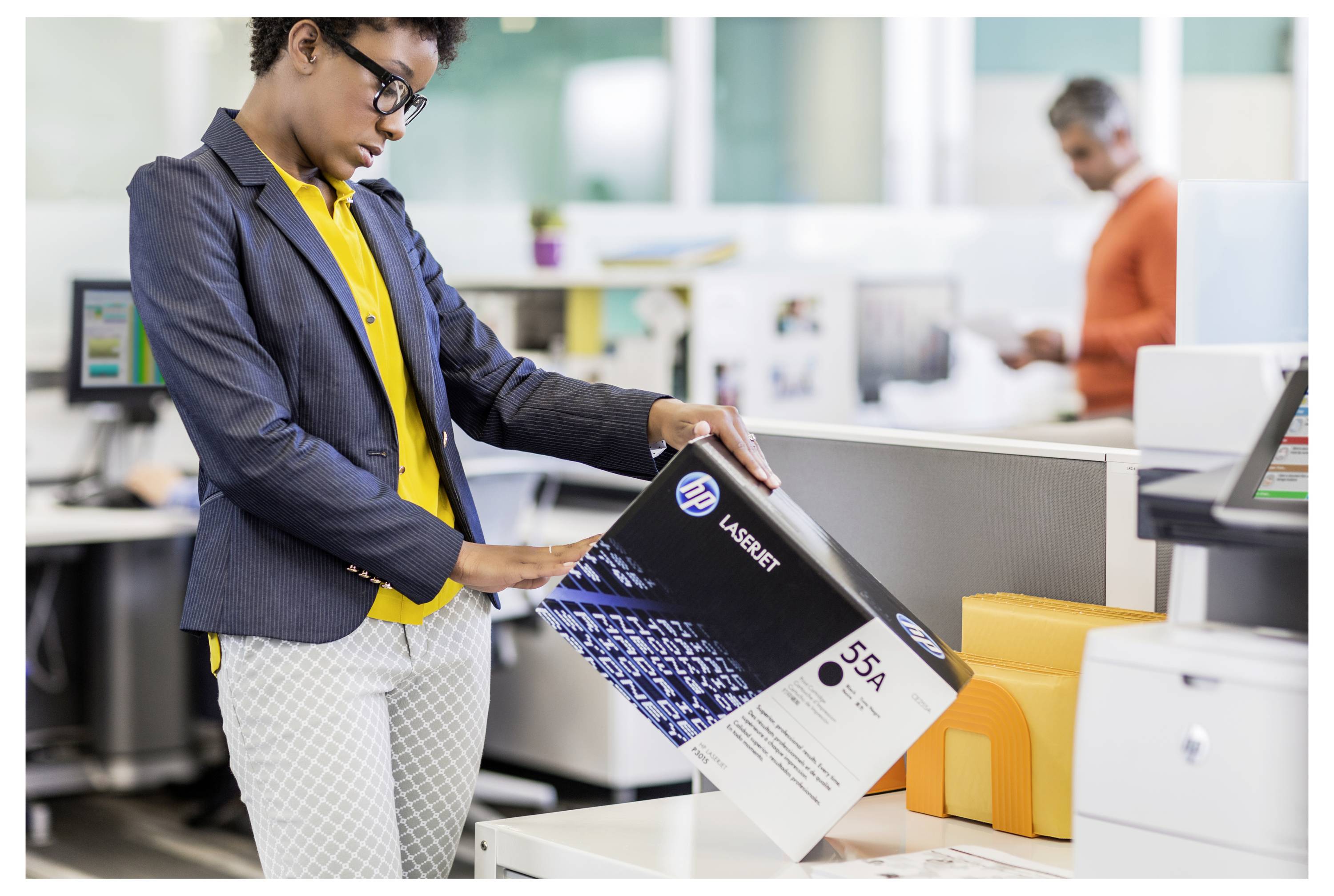 Vrouw in een kantoor selecteert een LaserJet 55A tonercartridge uit een stapel op het bureau, met een collega die in de achtergrond werkt.