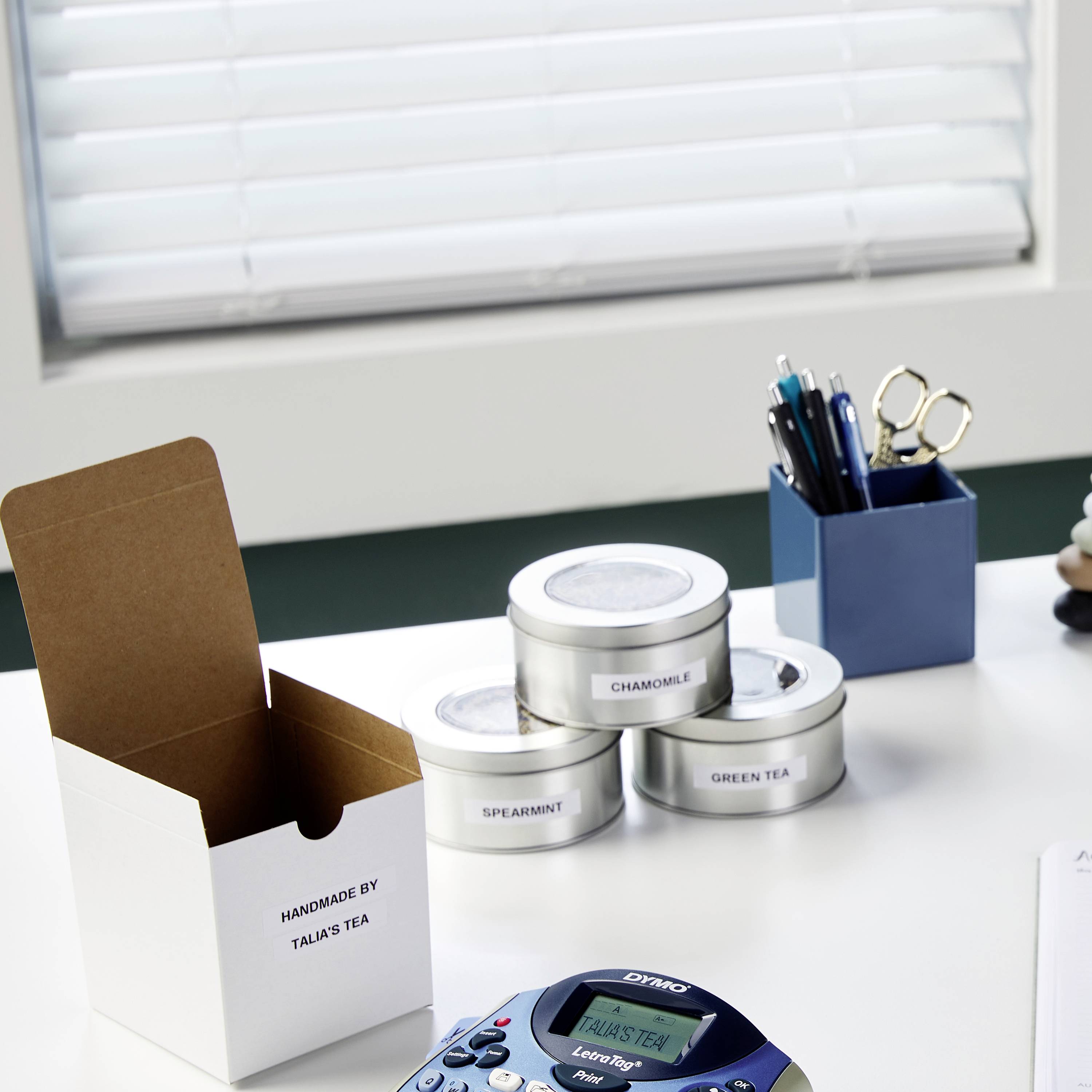 Een bureau met theedozen, een theebladtester, pennen in een houder en een kleine witte doos met de tekst 'Handmade by Talas Tea'.