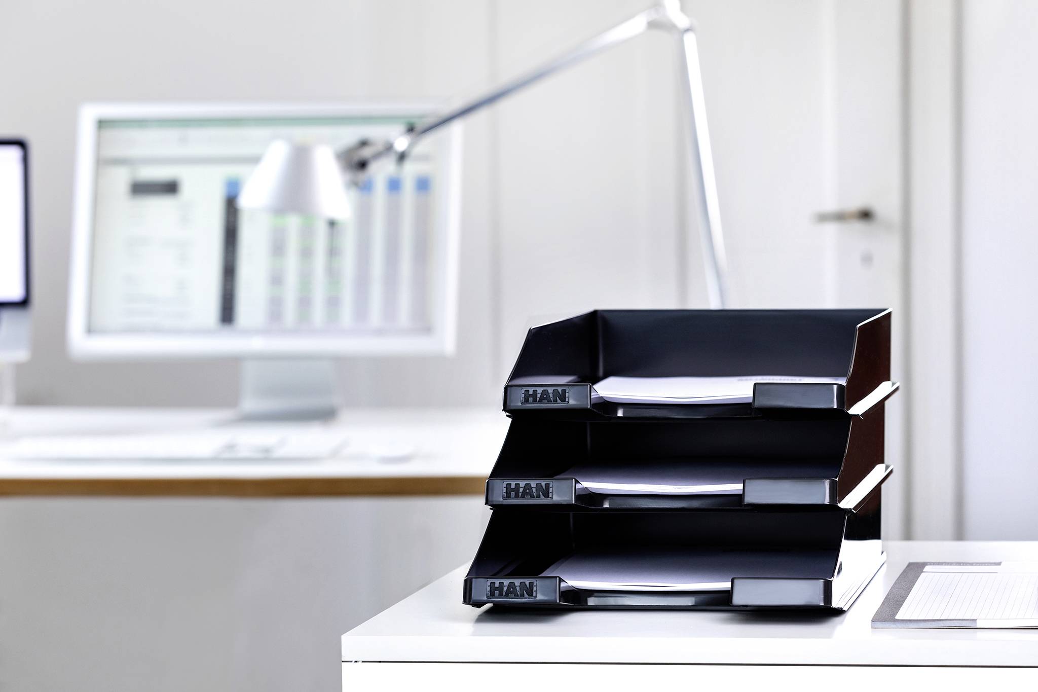 Een moderne werkplek met drie zwarte opbergvakken op een bureau, met op de achtergrond een monitor en een lamp.