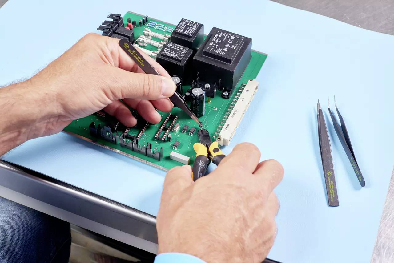 Handen repareren een printplaat met gereedschappen op een tafel, waarop elektronische componenten zoals chips en weerstanden liggen.