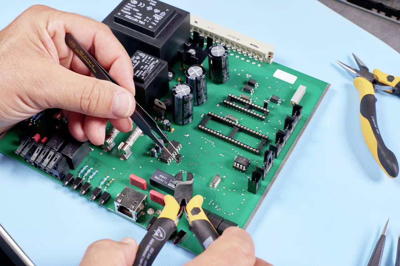 Een technicus repareert een printplaat met verschillende gereedschappen, waaronder een pincet en een zijkniptang.