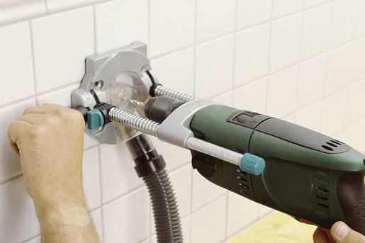 Een persoon boort met een boormachine een gat in een betegelde muur. Een stofzuiger verwijdert het ontstane stof.