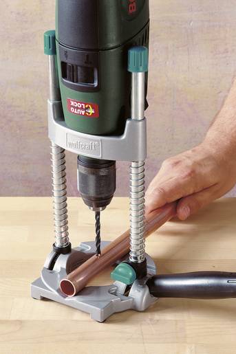 Een hand houdt een koperen buis vast op een tafel, terwijl een boormachine de buis doorboort.