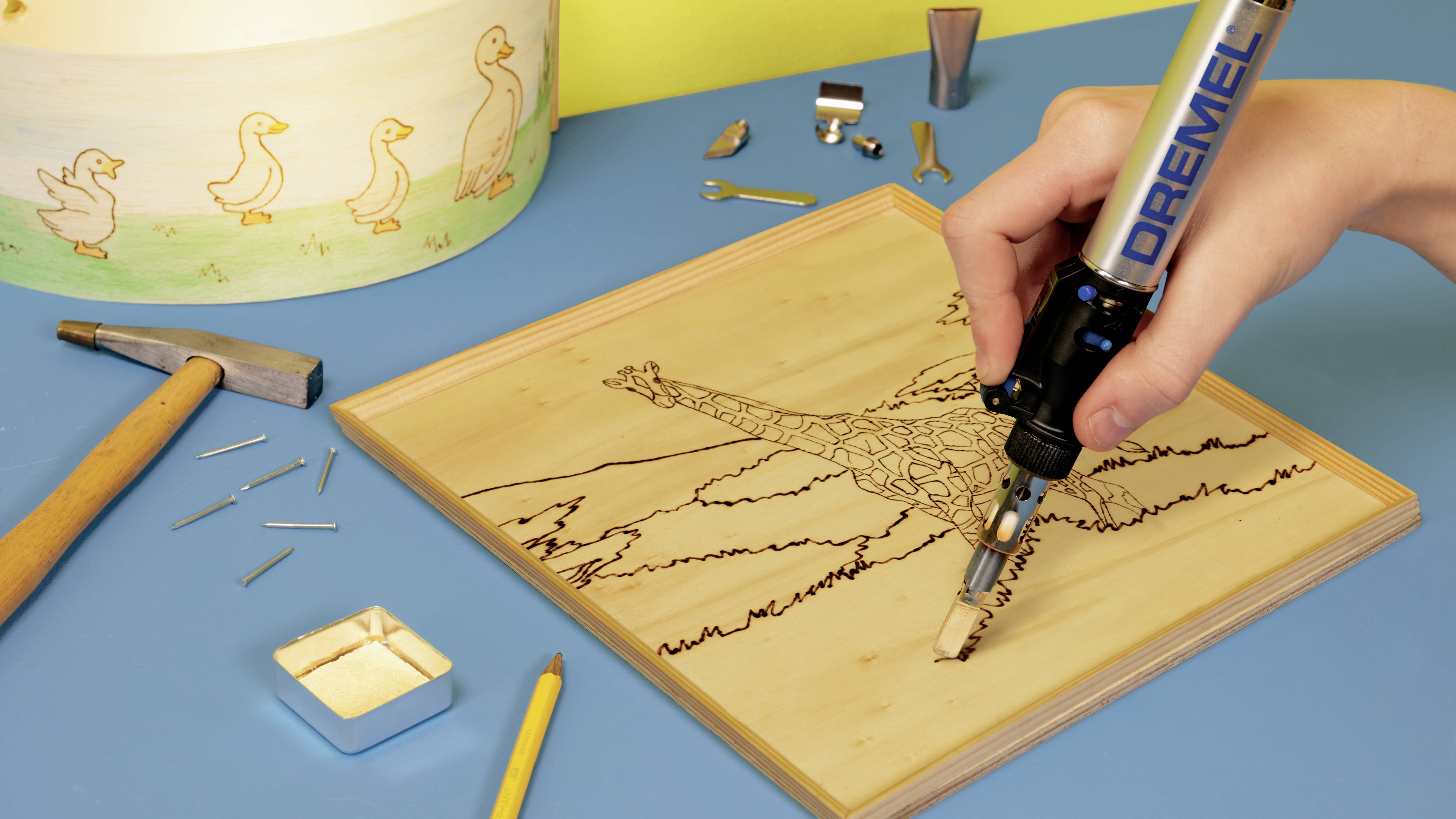 Een persoon brandt met een brandgereedschap het beeld van een giraf in een houten plaat. Op tafel liggen gereedschappen en spijkers.