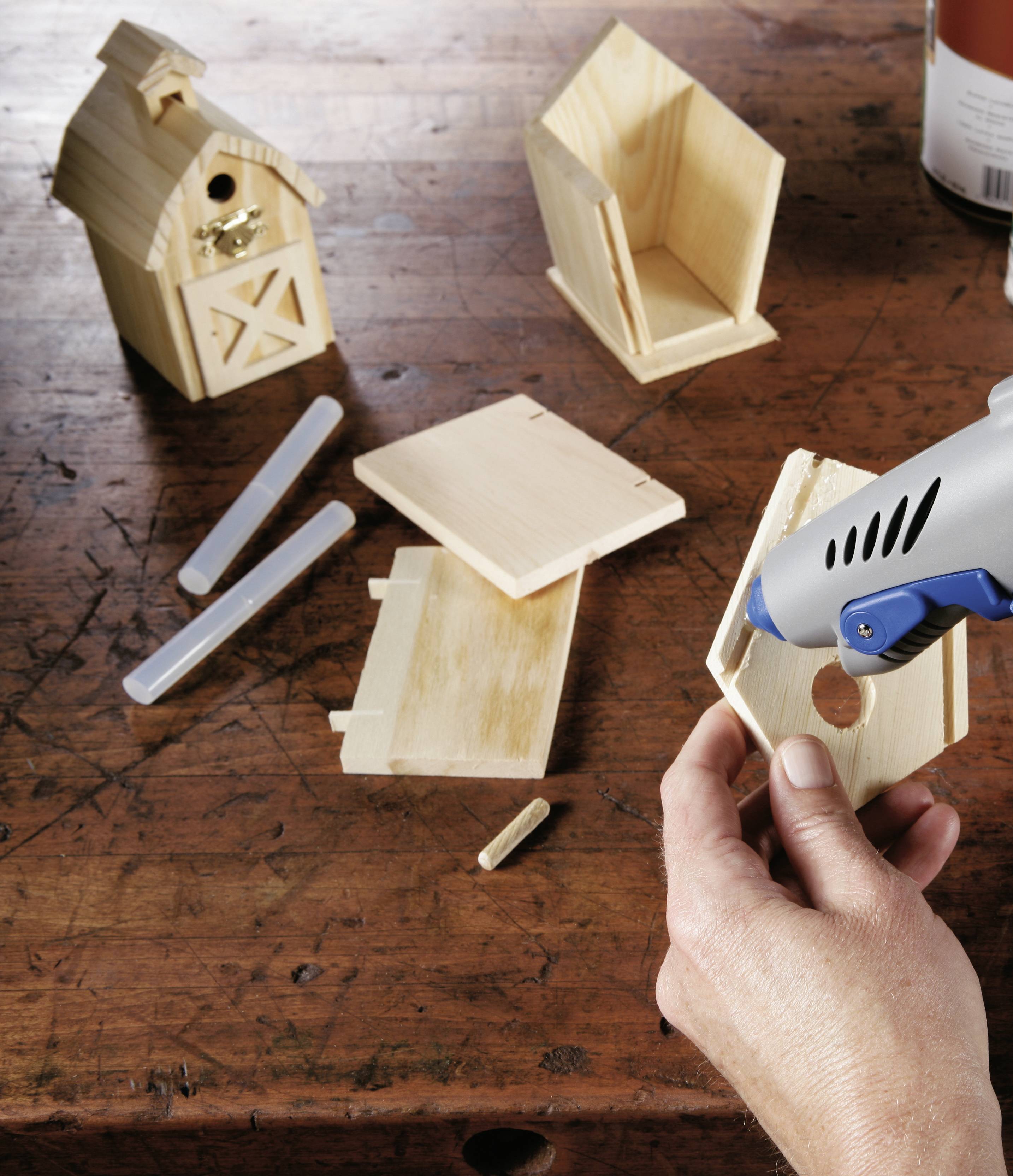 Een hand houdt een klein houten onderdeel vast dat wordt samengevoegd met een lijmpistool. Op de achtergrond zijn nog meer houten componenten zichtbaar.