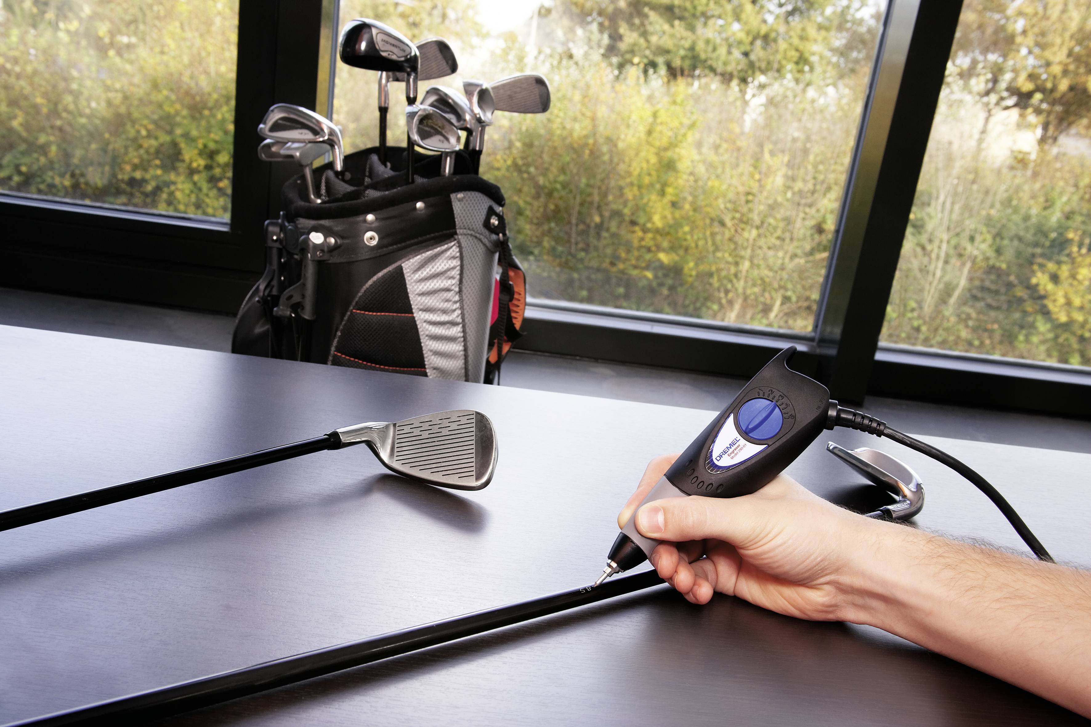 Een handapparaat meet de lengte van een golfclub op een tafel. Op de achtergrond staat een golftas met andere clubs.