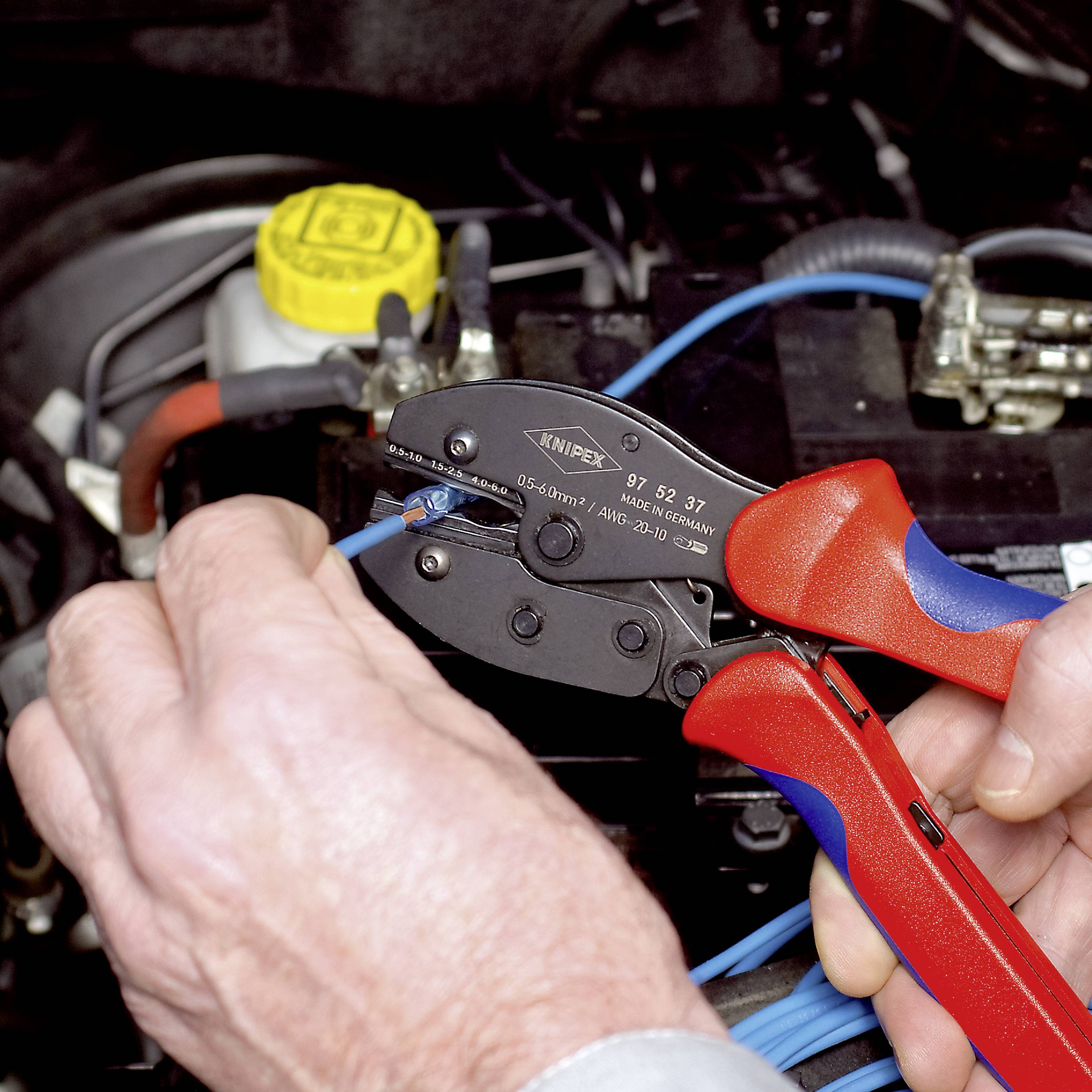 Een persoon gebruikt een rode en blauwe krimptang om elektrische bedrading te crimpen in een auto. Op de achtergrond is een motorruimte zichtbaar.