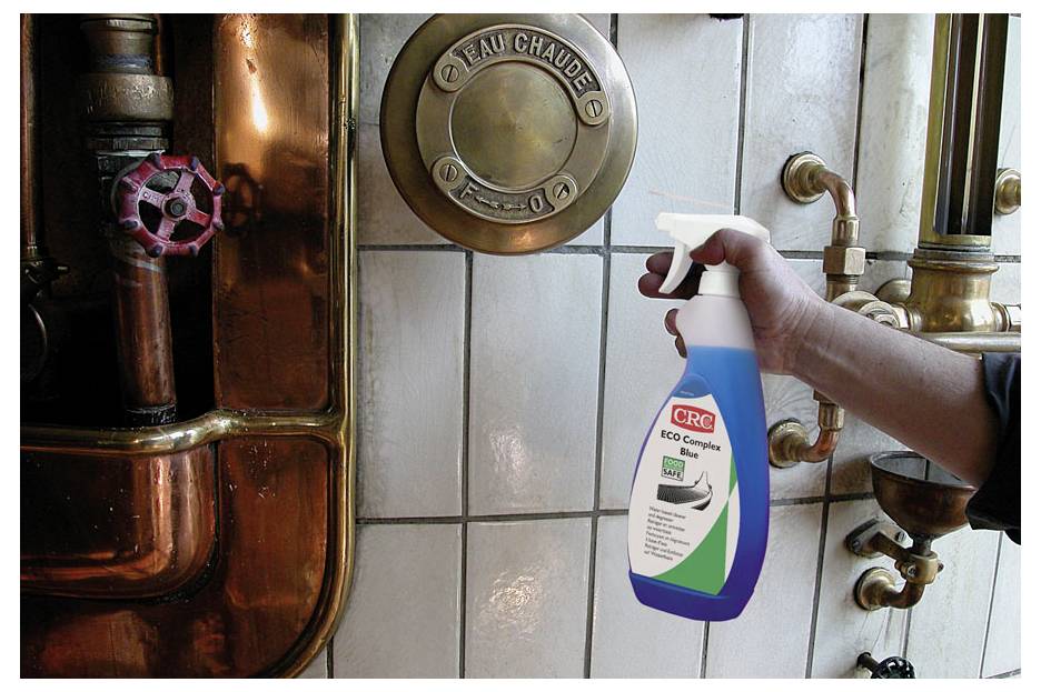 Een hand houdt een CRC ECO complex reinigingsspray bij industriële leidingen, wat wijst op het gebruik ervan voor het reinigen van buizen of apparatuur.