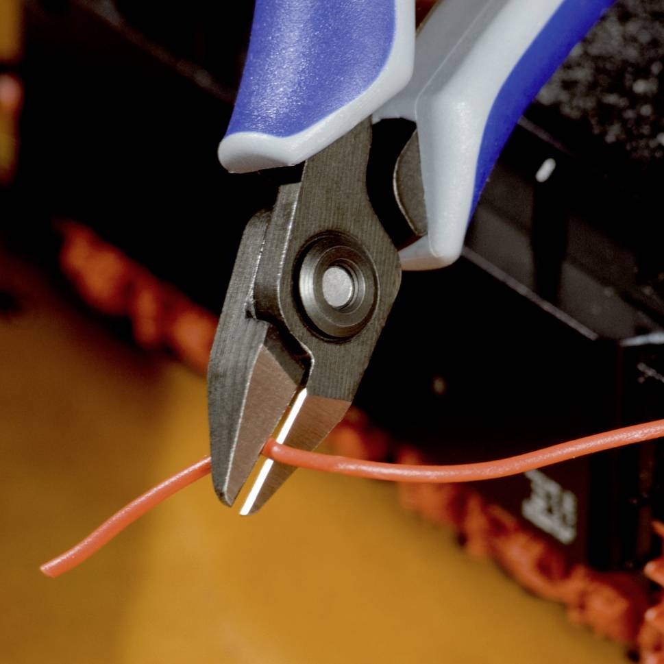 Close-up van een tang die een rood kabeltje doorsnijdt. Scherpe focus op de knipactie, op de achtergrond onscherpe onderdelen van een werkbank te zien.