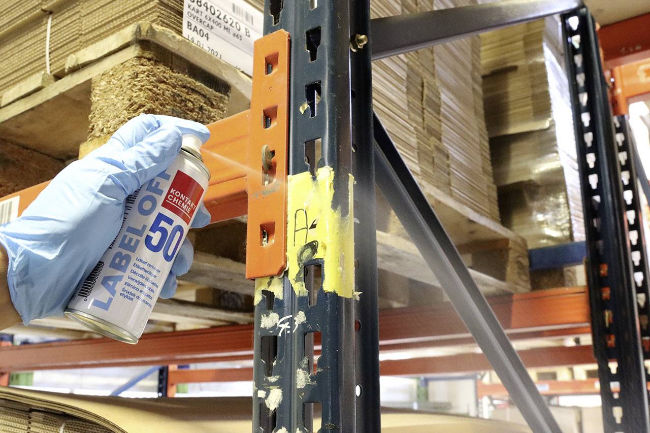 Een handschoendragende persoon houdt een blik etikettenverwijderaar vast bij een rek in een magazijn, om etikettenresten van de metalen zuil te verwijderen.