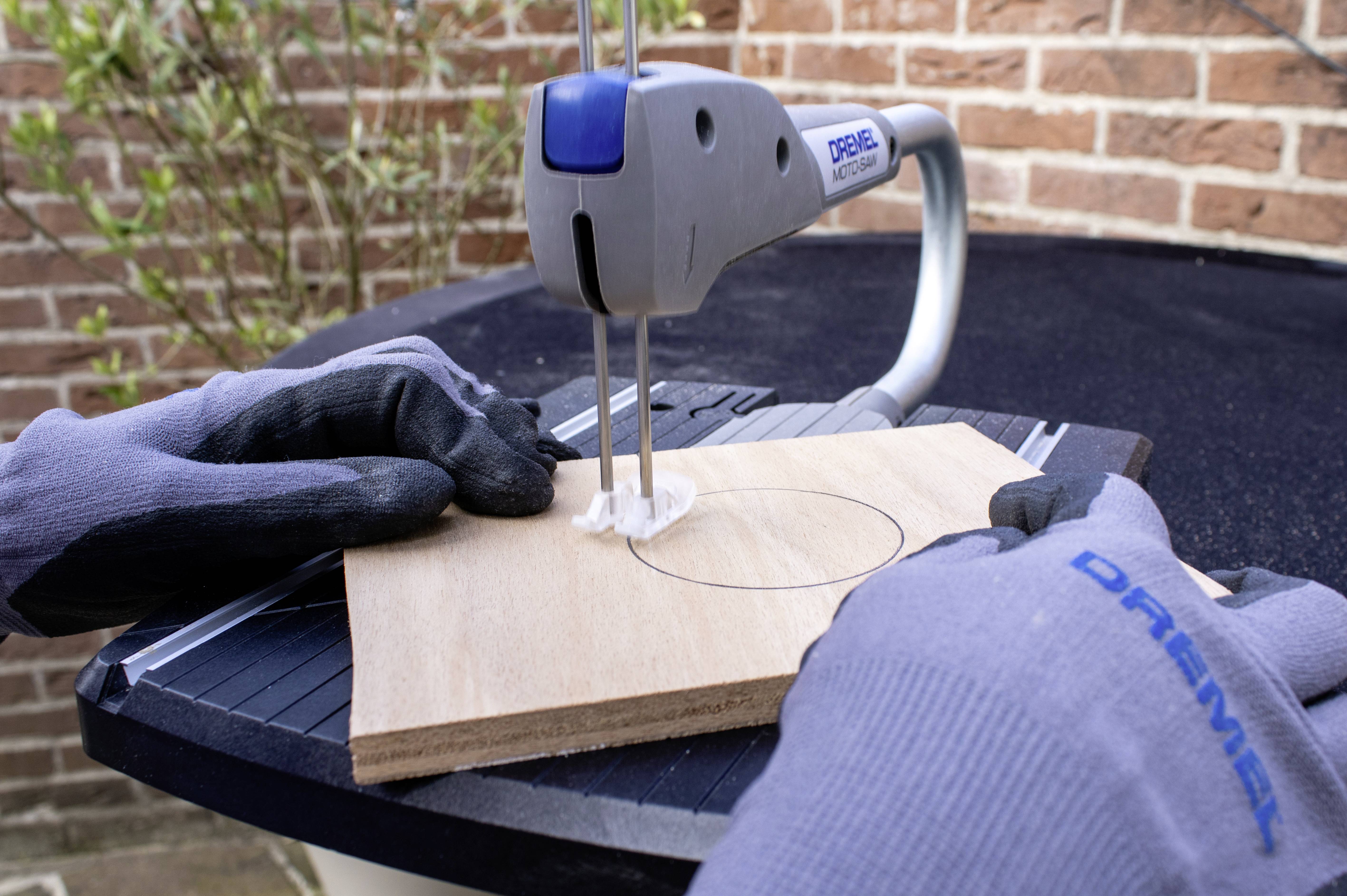 Een persoon zaagt een rond stuk hout uit met een elektrische decoupeerzaag. Veiligheidshandschoenen worden gedragen.