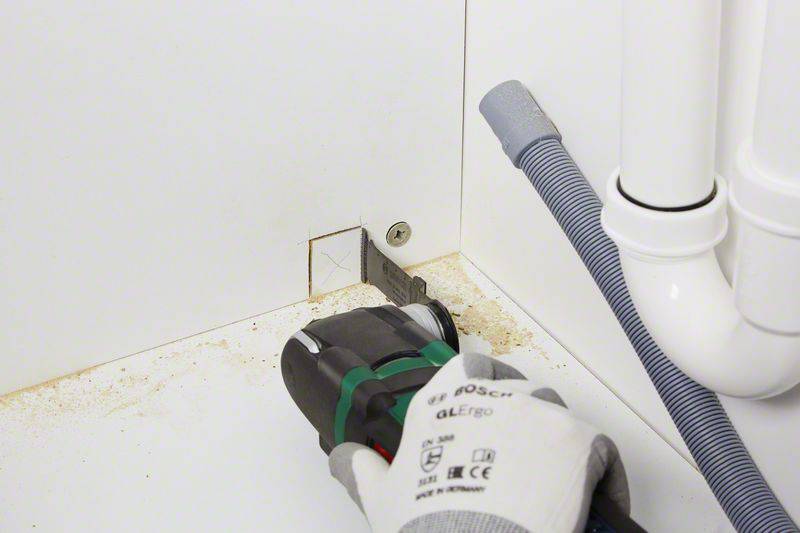 Een elektrisch gereedschap wordt gebruikt om een vierkant gat te maken in een witte wandplaat onder een wastafel.