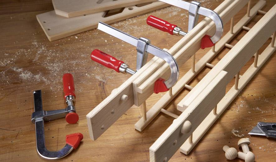 Houten onderdelen worden met lijmklemmen op een werkbank vastgezet. Gereedschappen en zaagsel liggen verspreid, wat ambachtelijk werk illustreert.