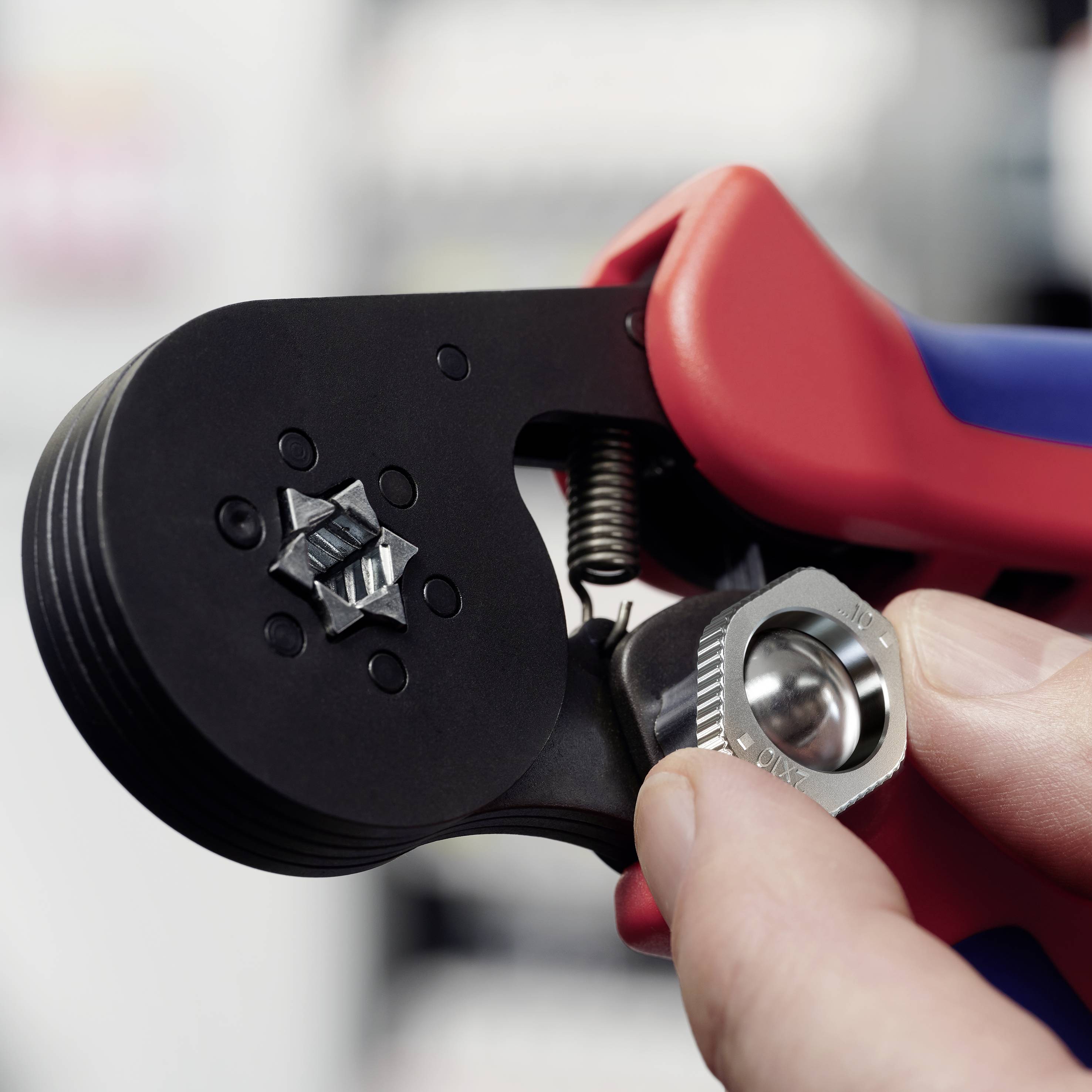 Een hand houdt een krimptang vast die een elektrische kabel knelt. De tang is zwart en rood, de achtergrond is onscherp.