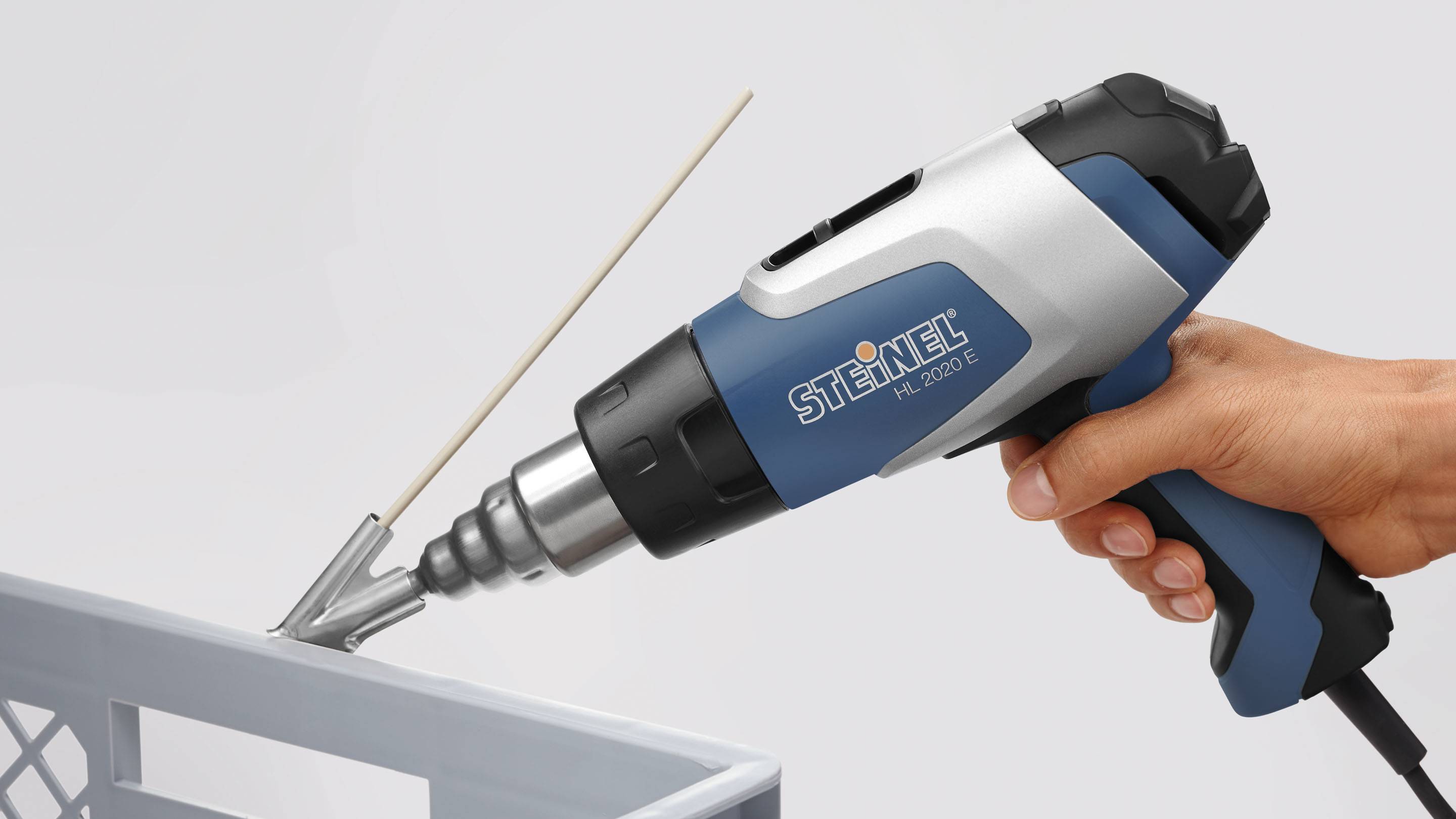 Een hand houdt een heteluchtpistool vast met het opschrift 'Steinel HL 2020 E', gericht op een grijze kunststofoppervlakte.