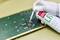 Hand met een witte handschoen sproeit een printplaat met een reinigingsspray. De printplaat is bedekt met groene en gouden componenten.