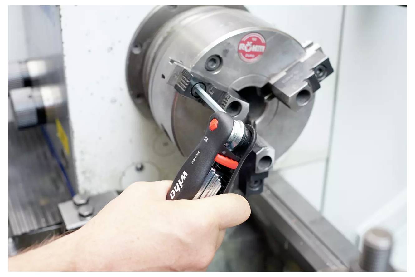 Een hand gebruikt een zeskantige sleutel om een metalen onderdeel op een draaibank in een werkplaatsomgeving bij te stellen.