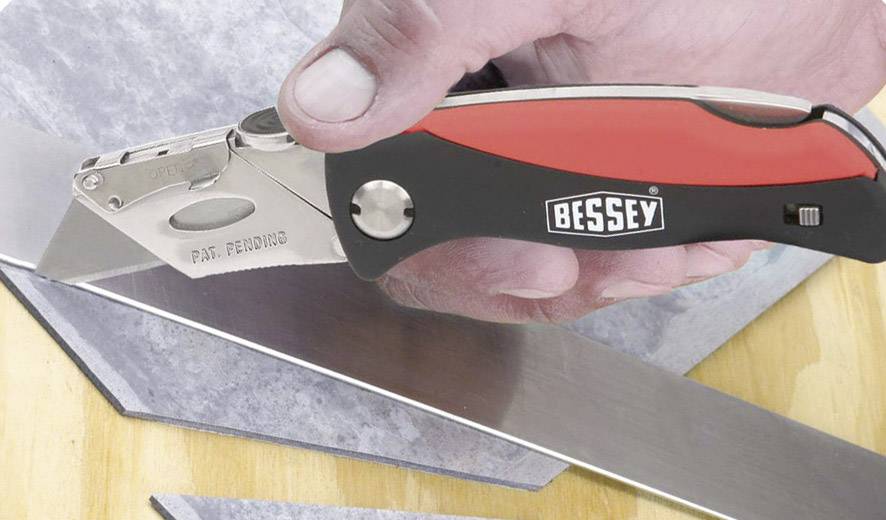 Een hand houdt een tapijtmes met een rode handgreep van het merk Bessey en snijdt een grijze oppervlakte.
