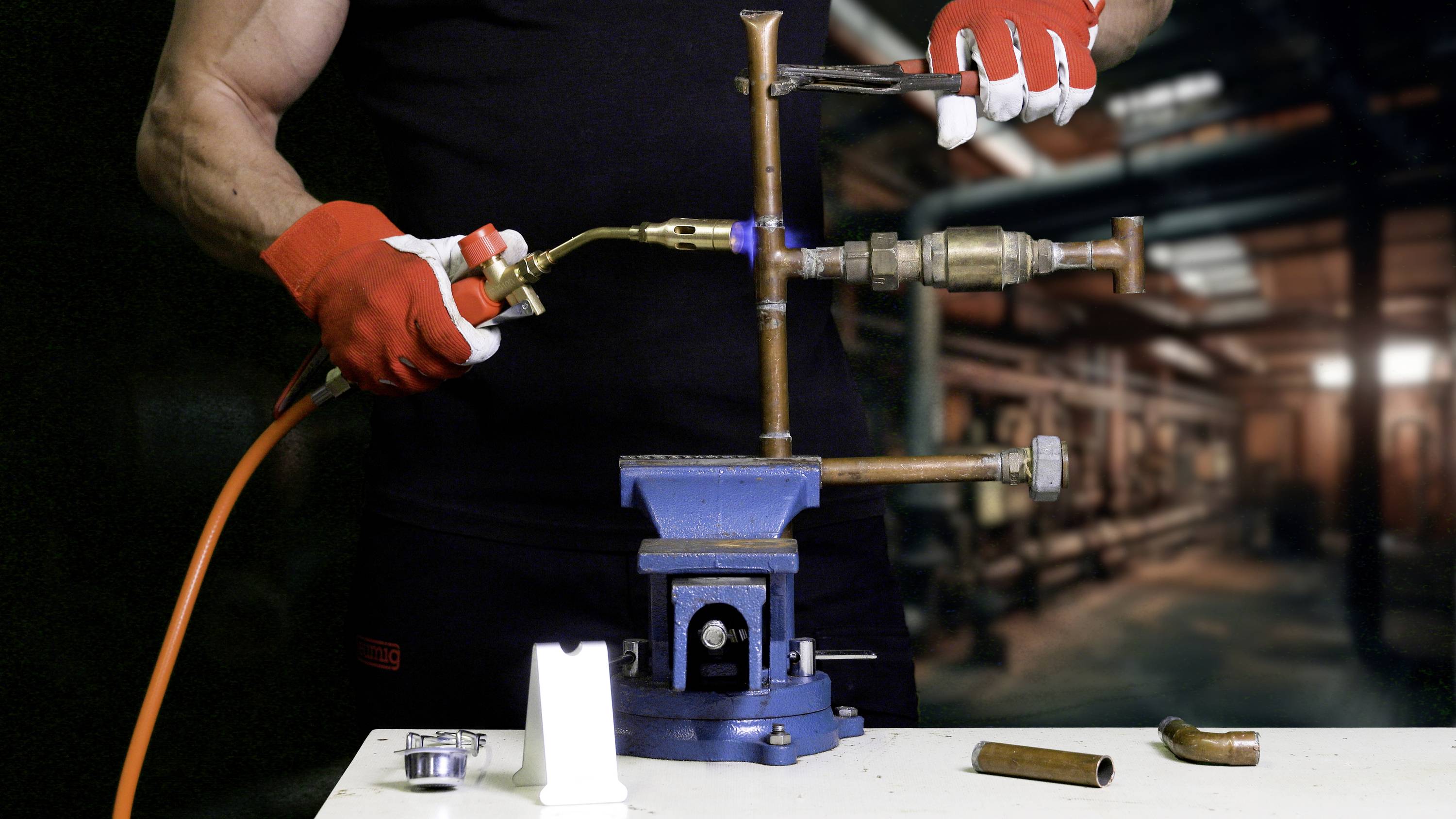 Een persoon met rode handschoenen soldeert koperen buizen in een werkplaats. Een bankschroef houdt de buizen vast, en een soldeerlamp wordt gebruikt.