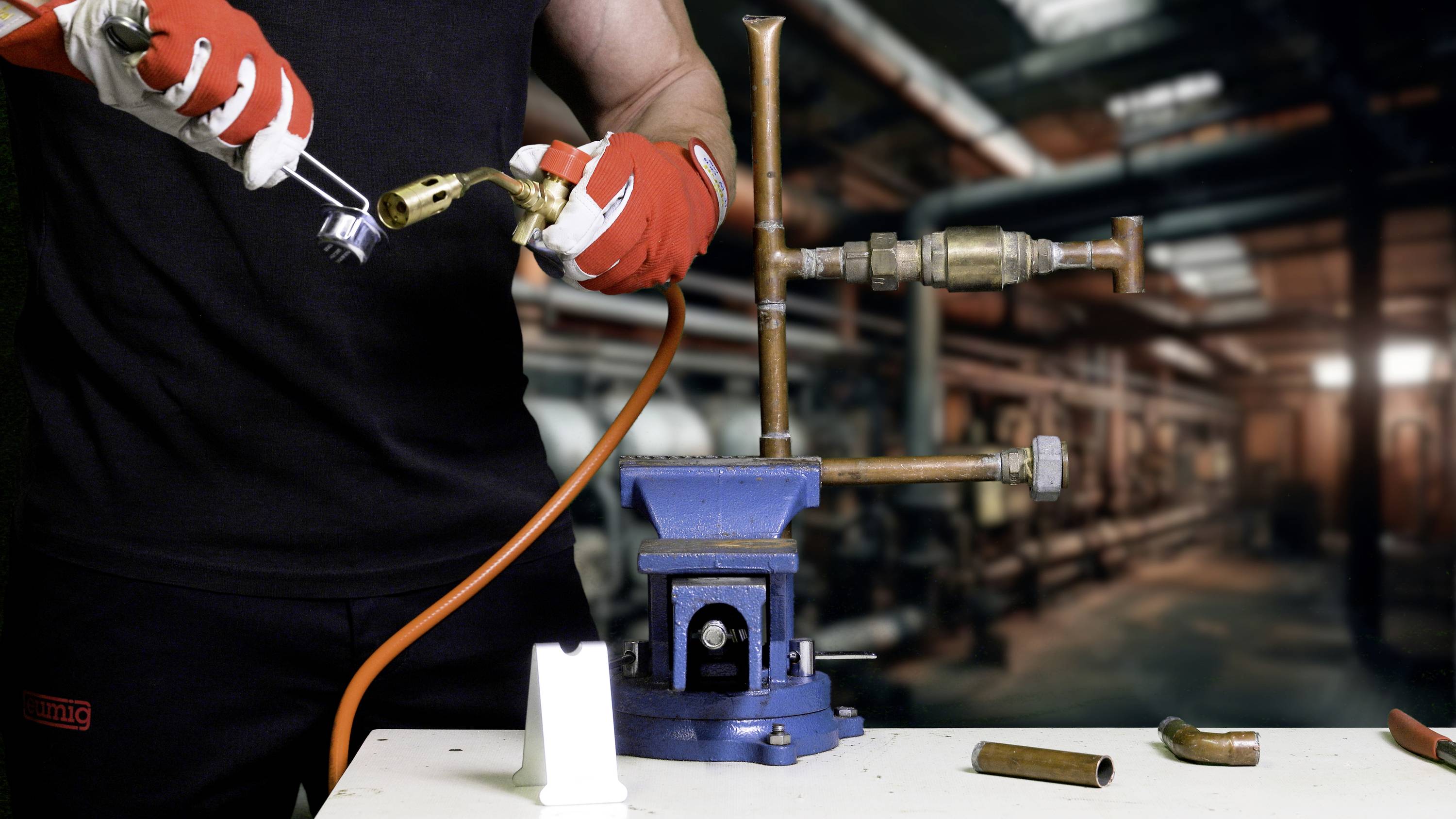 Een persoon in beschermende kleding werkt met metalen buizen in een werkplaats. Een bankschroef houdt de buizen vast terwijl de arbeider ze bewerkt.