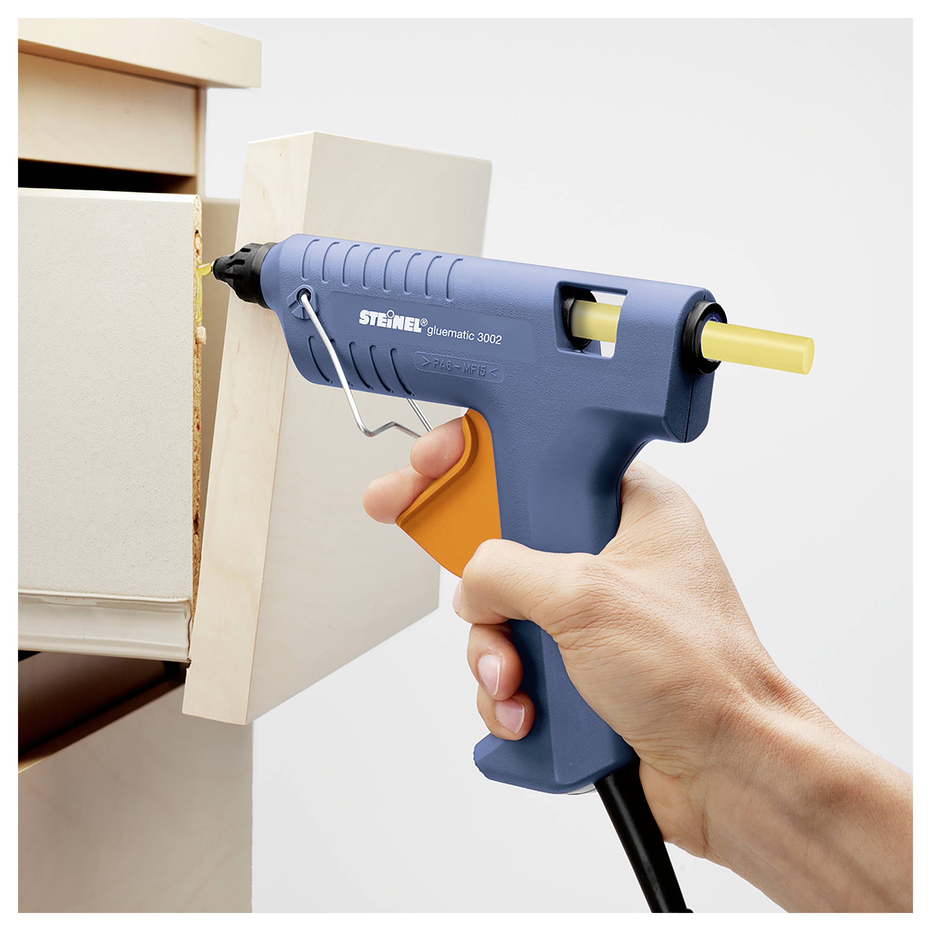 Hand die een blauwe hete lijmpistool gebruikt om lijm aan te brengen op een houten paneel op een licht gekleurde kast of doos.