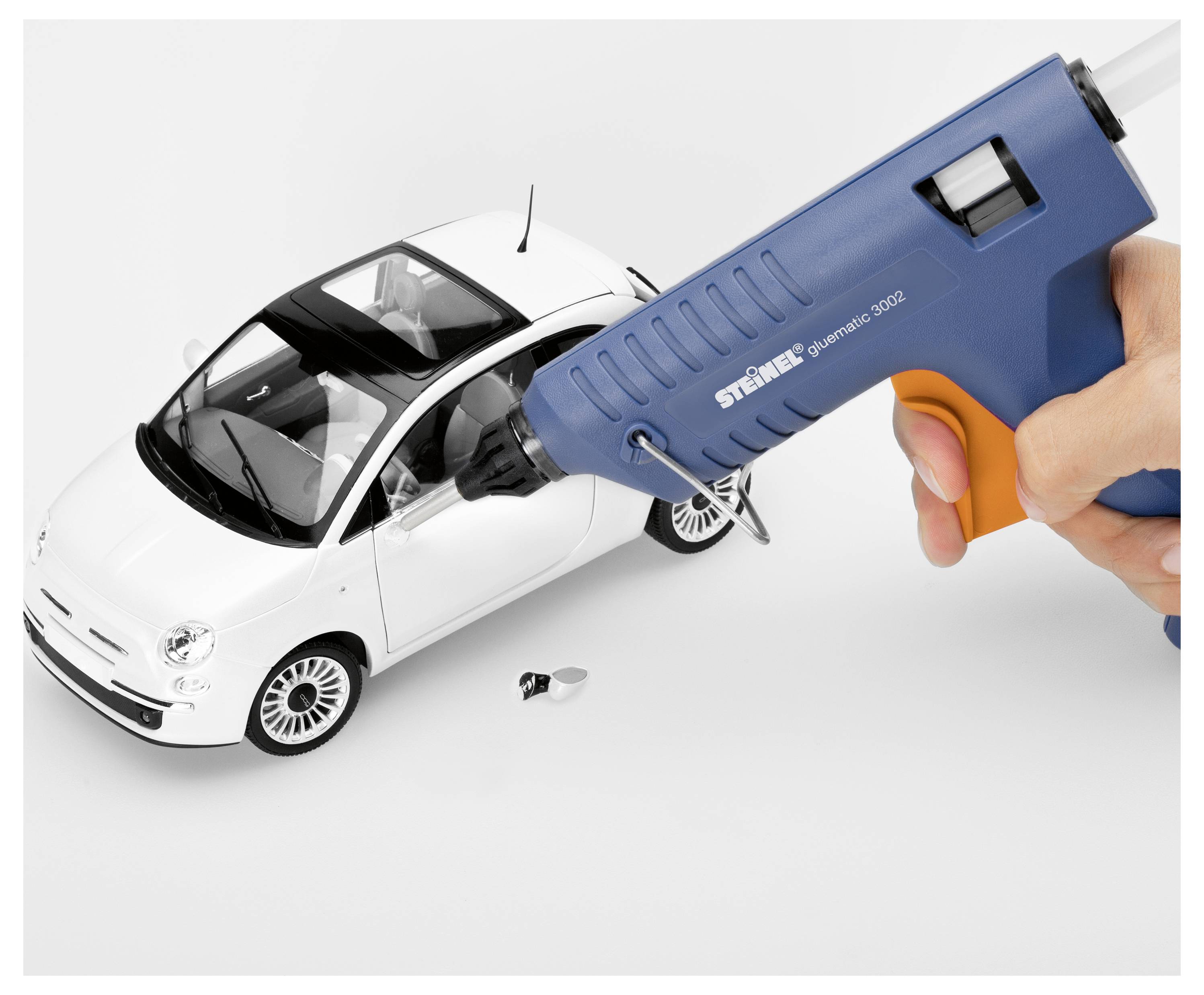 Hand die een blauwe hete lijmpistool vasthoudt, brengt lijm aan op een wit speelgoedautomodel op een effen achtergrond.