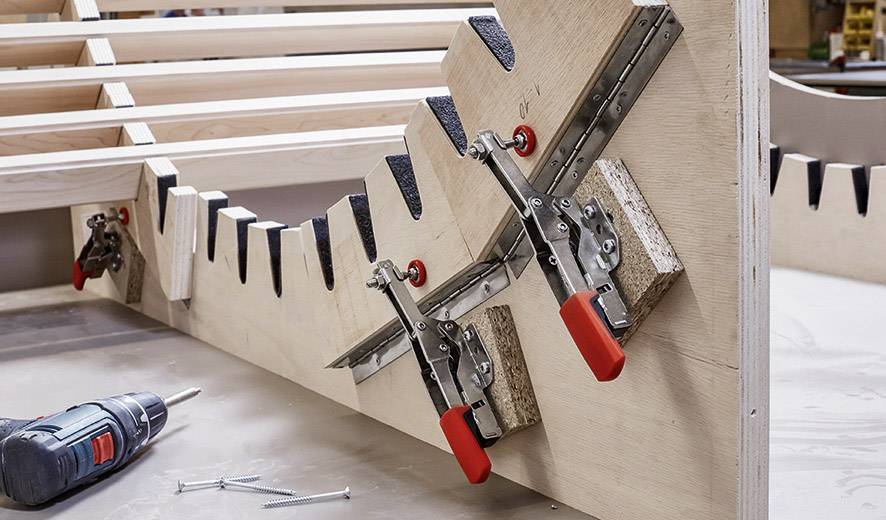 Houten stuk wordt met klemmen vastgezet om een gebogen constructie te creëren. Naast de boormachine en schroeven op de werkbank.