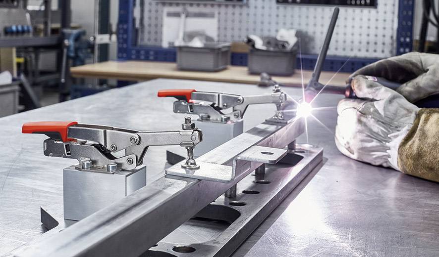 Lasstafel in een werkplaats, waarop een metalen onderdeel met klemmen is bevestigd. Een lichtstraal toont het lasproces.