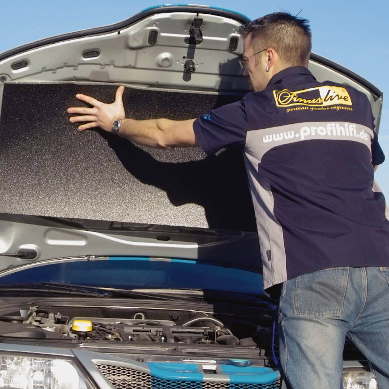 Een man staat voor een auto met een open motorkap en bevestigt een geluidsisoleringmat aan de binnenkant van de motorkap. De man draagt een poloshirt met een opdruk op de rug.