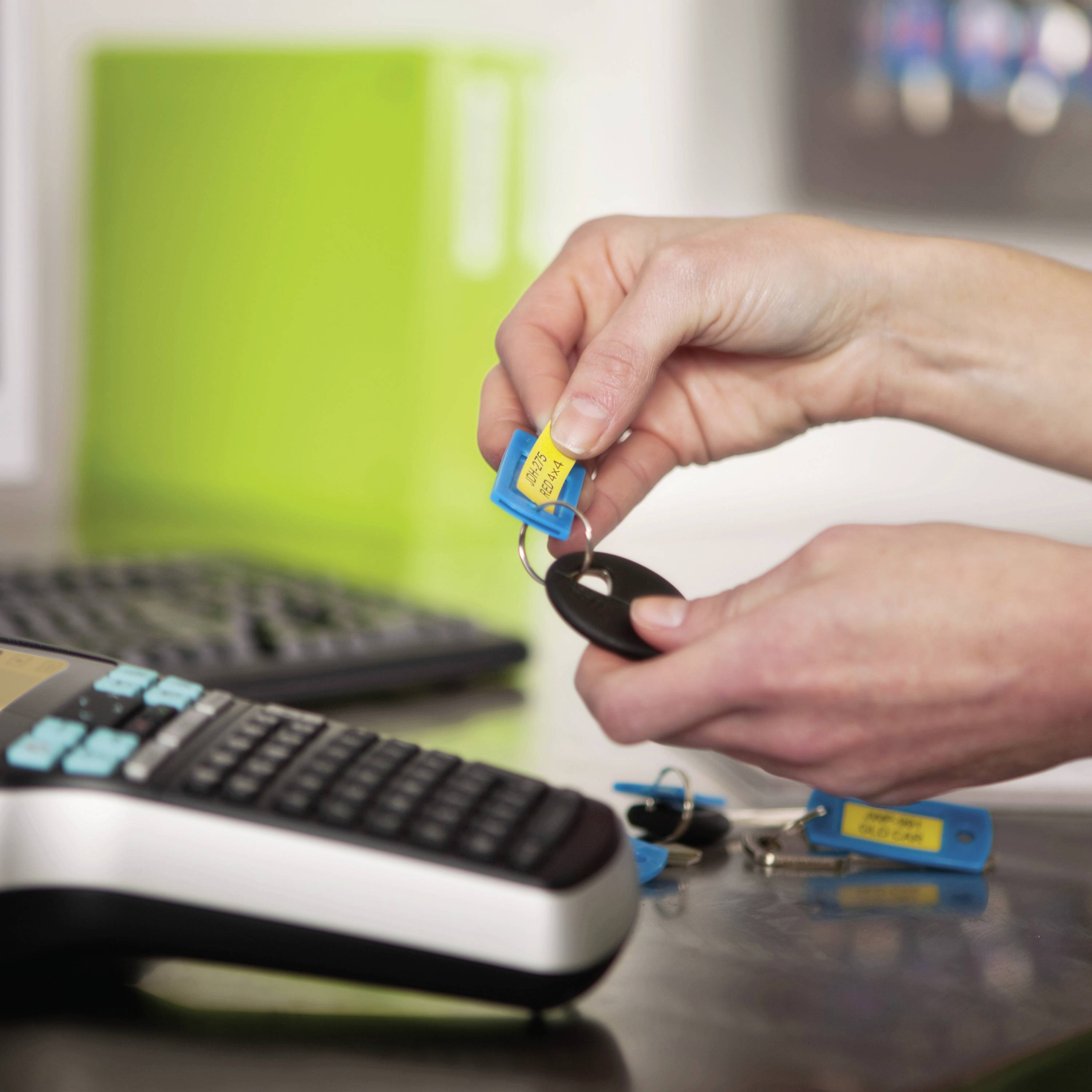 Een hand houdt een blauwe sleutelhanger met tekst vast. Ervoor ligt een toetsenbord. Op de achtergrond staat een groene map.