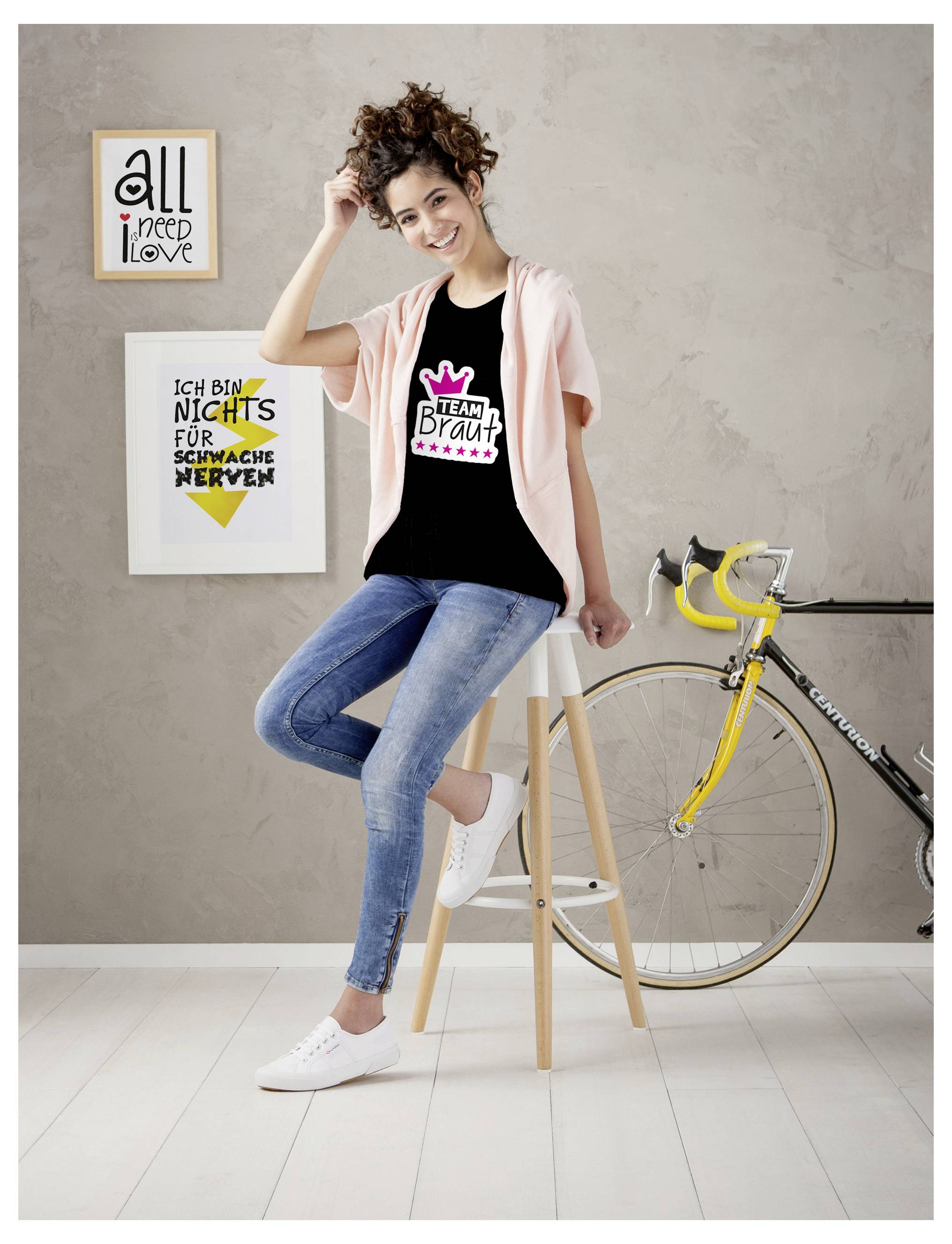 Jonge vrouw zit glimlachend op een kruk naast een fiets; haar T-shirt draagt de opdruk 'TEAM BRUID'. Aan de muur hangen twee posters.