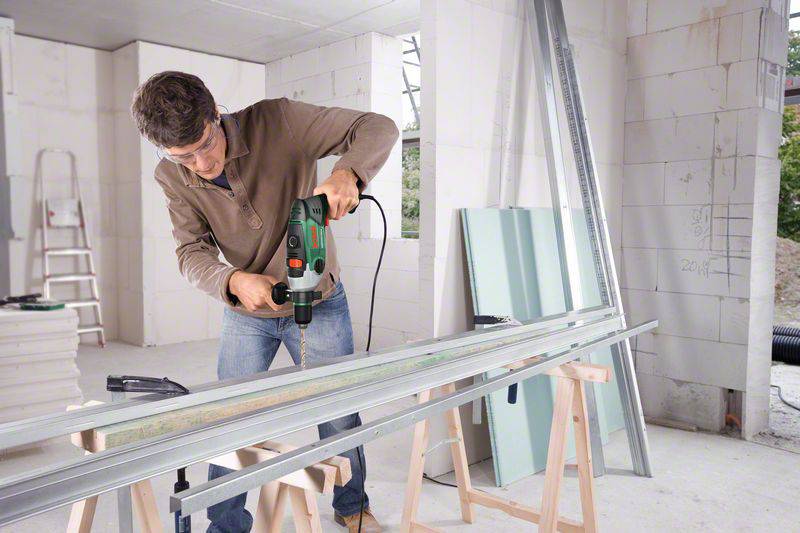 Een man met veiligheidsbril gebruikt een boormachine bij het bouwen van een metalen frame in een onafgewerkte ruimte. Op de achtergrond zijn bouwmaterialen zichtbaar.