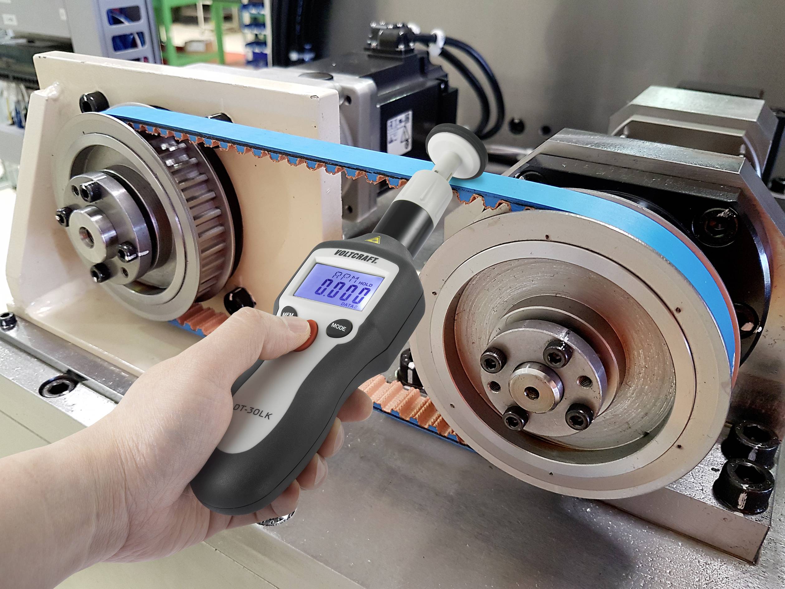 Een hand houdt een digitaal apparaat vast dat de spanning meet van een blauw-grijze riem tussen twee grote tandwielen.