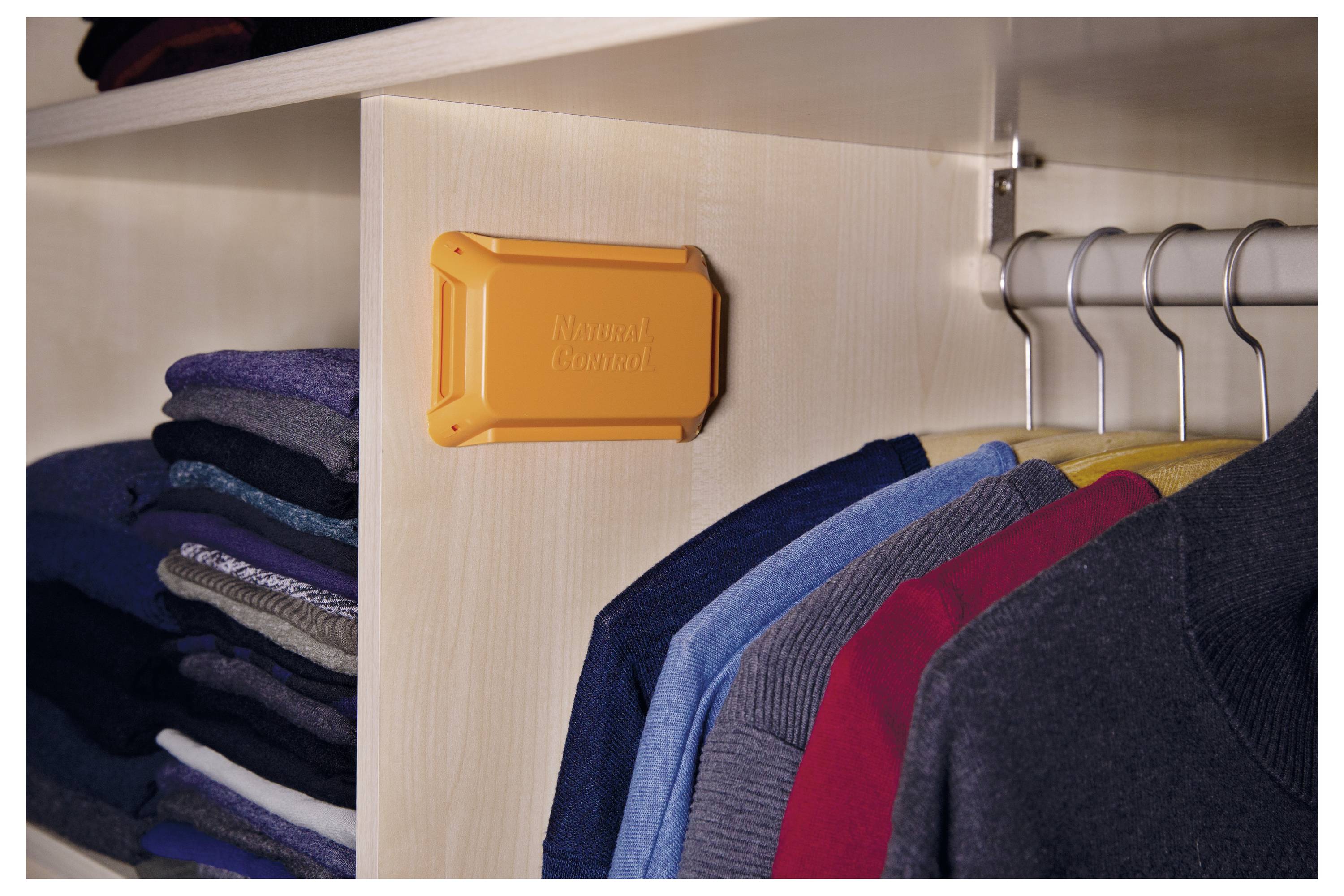 Kleren die hangen in een kledingkast met een oranje plastic apparaat dat aan de zijkant is bevestigd. Op het apparaat staat 'Mottenbestrijding'.