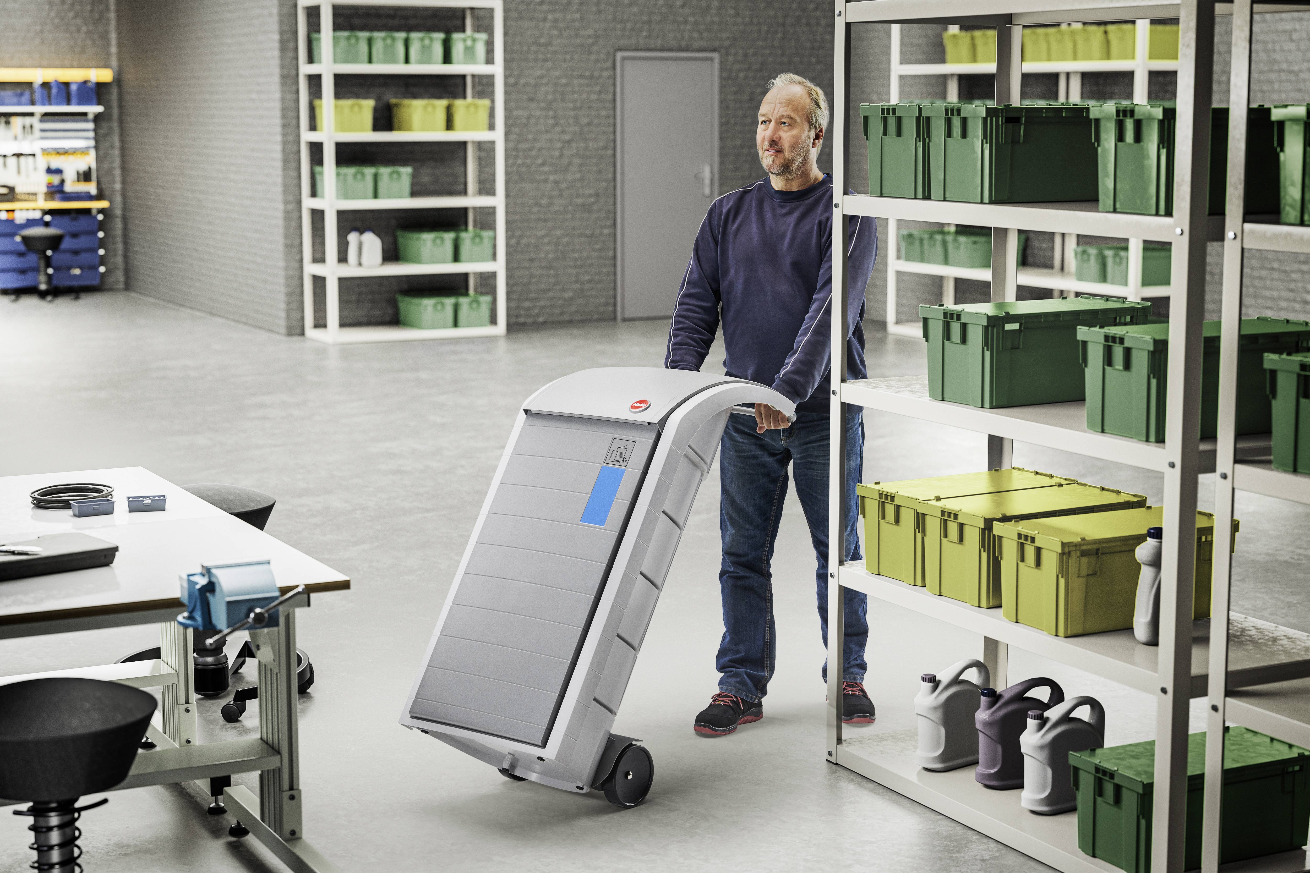 Een man duwt een grote mobiele container door een opslagruimte. In de ruimte staan stellingen met groene en gele dozen.