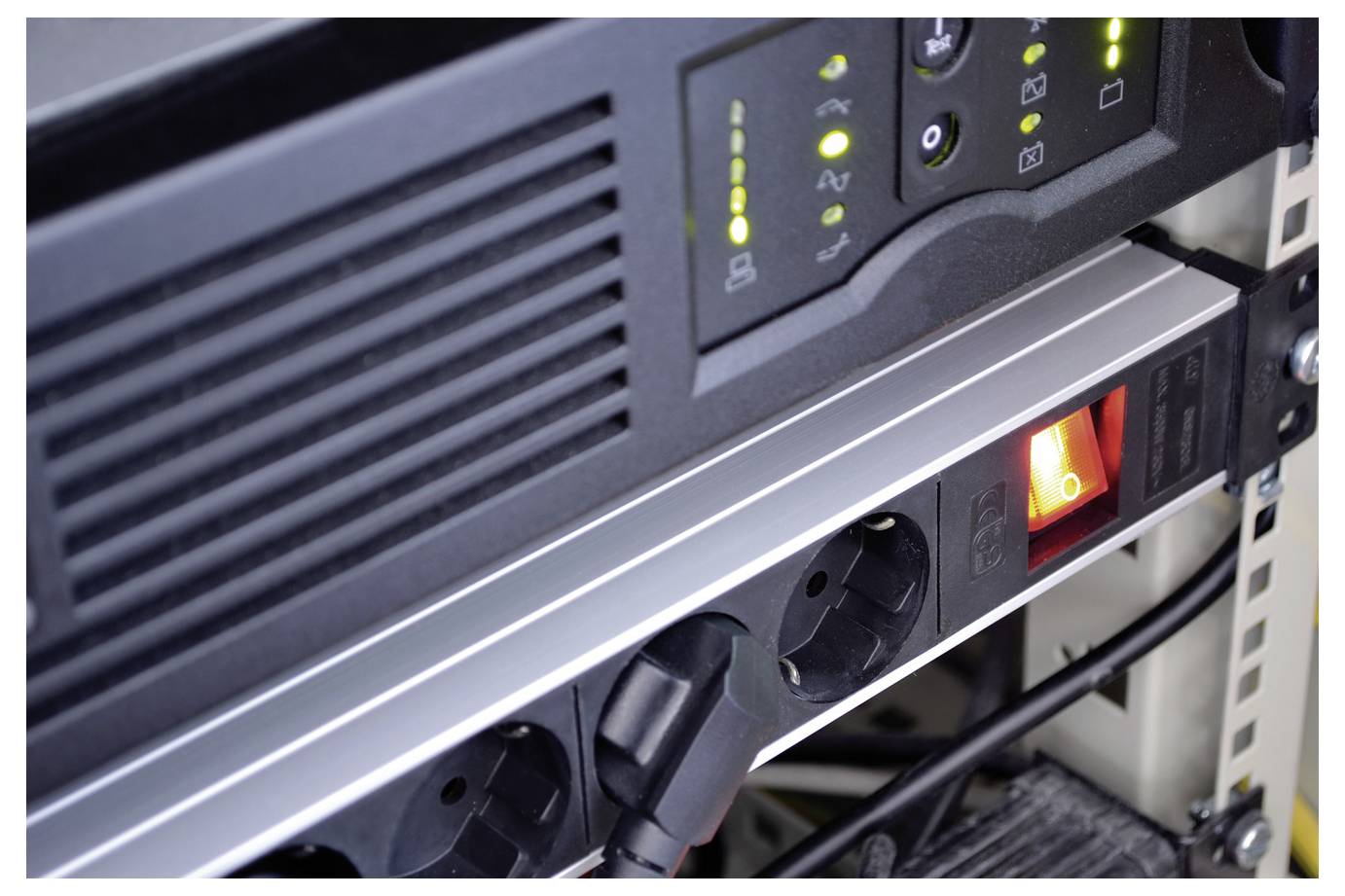 Close-up van een serverkaststroomstrip met verlichte schakelaar en meerdere ingestekerde snoeren, waarop aangeschakelde apparatuur zichtbaar is.