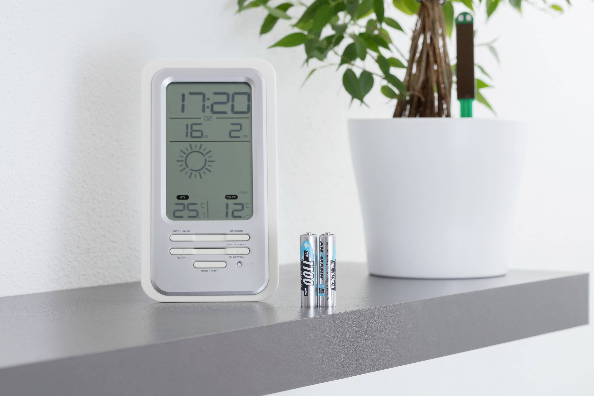 Een digitale thermo-hygrometer toont de tijd 17:20, temperatuur 25,1°C, luchtvochtigheid 12%, staat op een plank naast een bloempot.