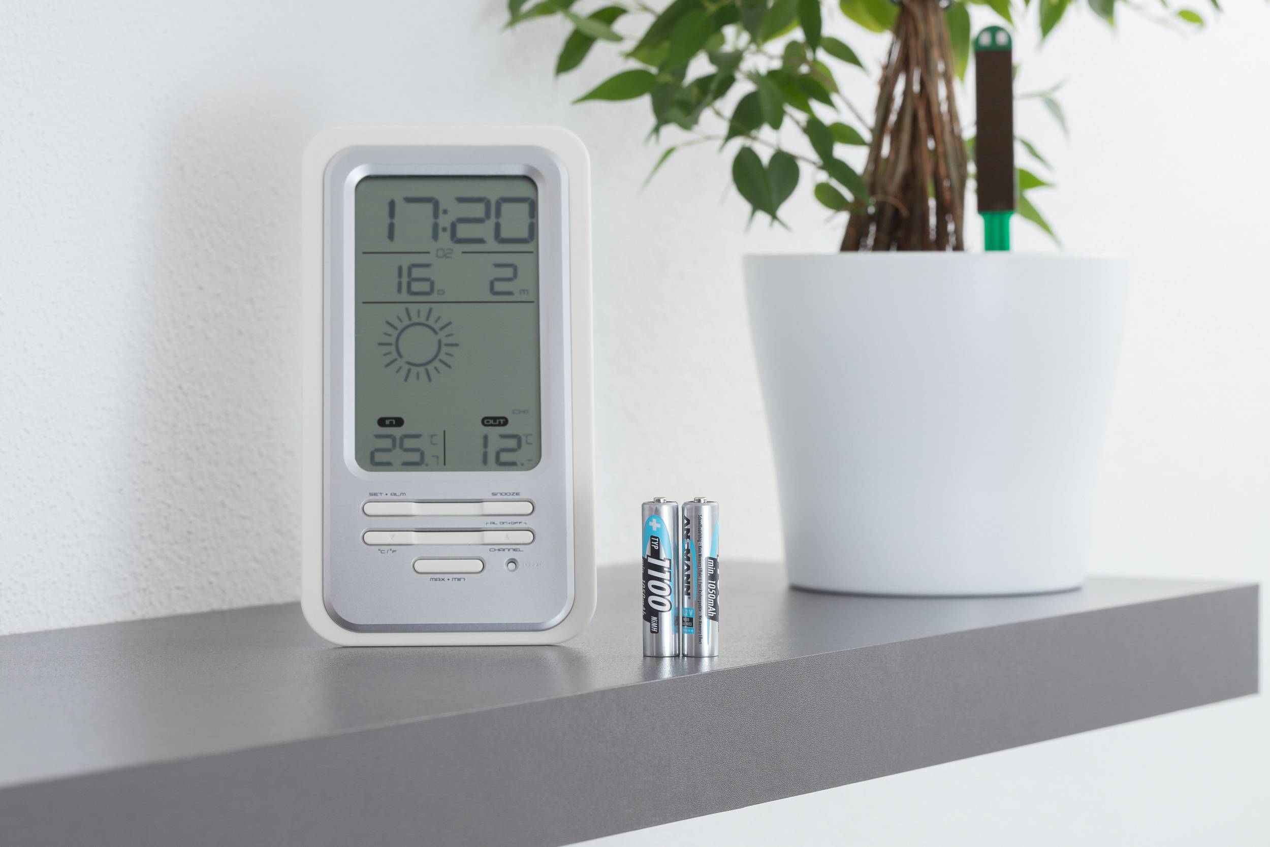 Digitale thermometer toont 17:20 uur, 25,1°C, zonnesymbool. Twee batterijen ervoor, bloempot op de achtergrond.