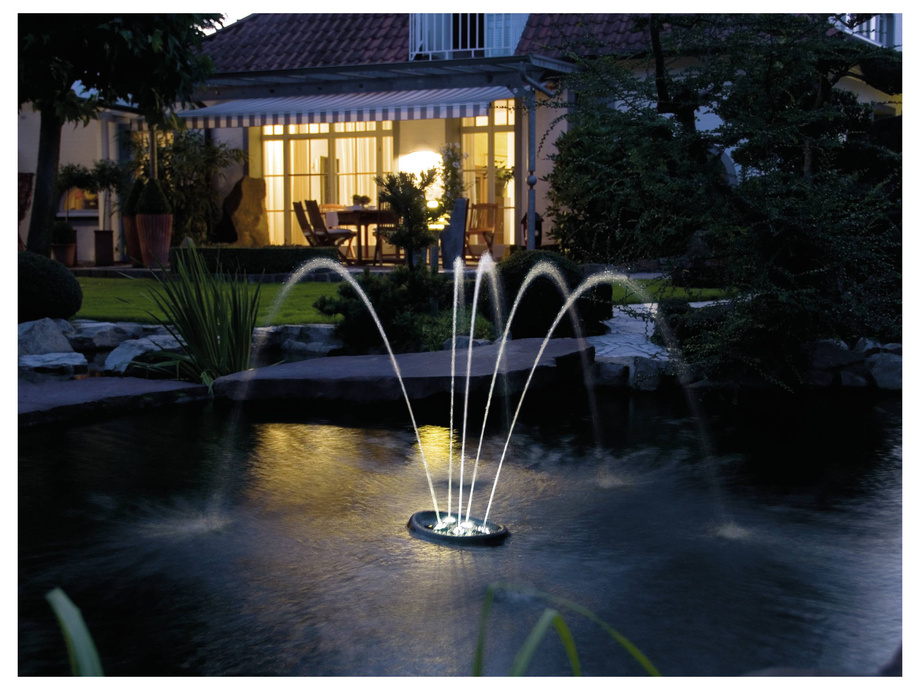 Een verlichte fontein in een tuinvijver 's nachts, met een warmverlichte binnenruimte van een huis zichtbaar op de achtergrond, wat een serene sfeer creëert.