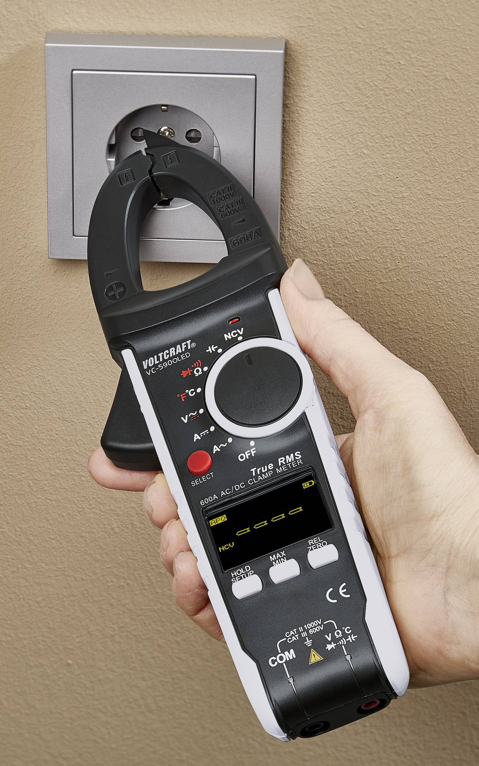 Een hand houdt een multimeter met tang vast bij een Europees stopcontact om de stroom te meten.