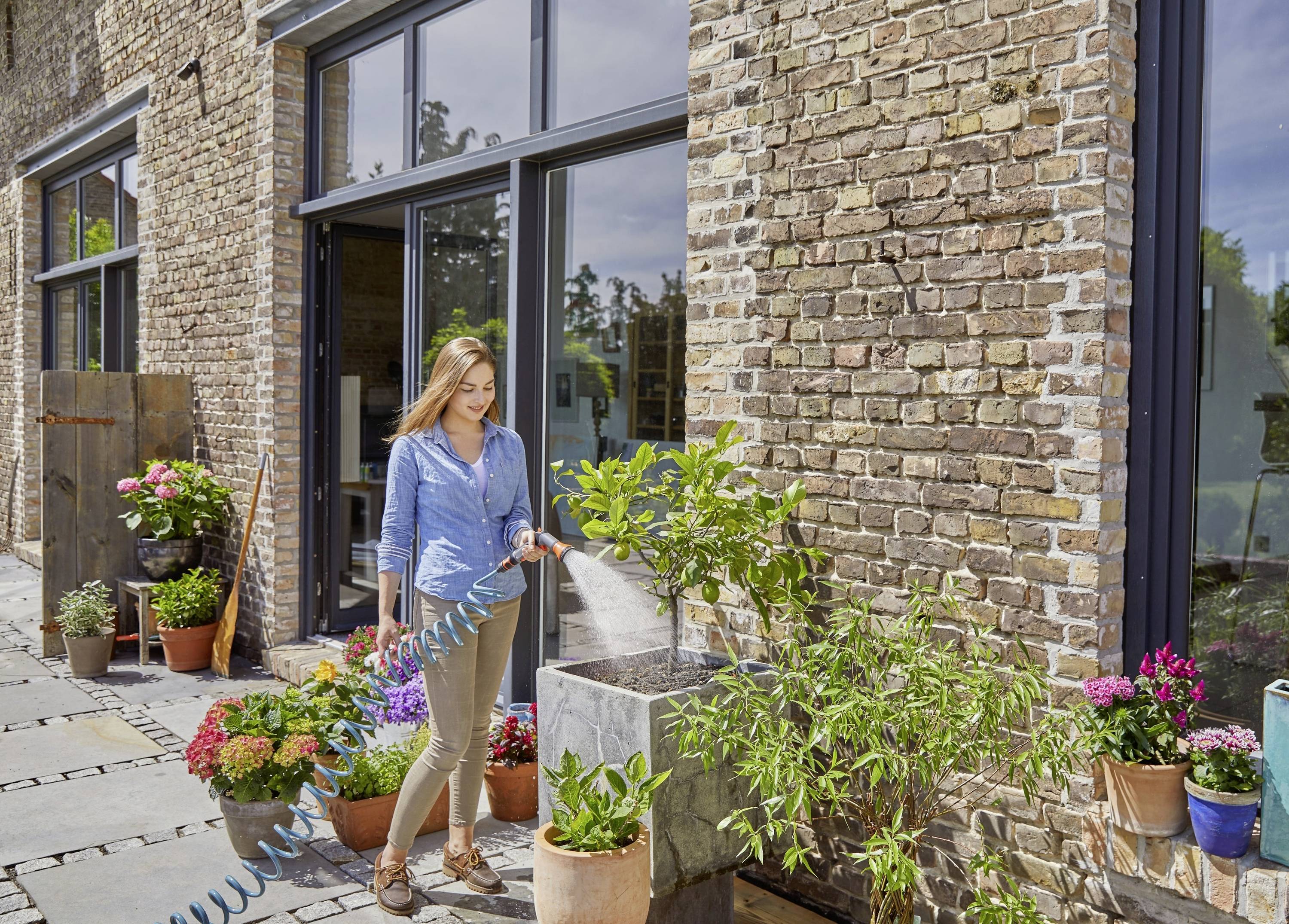 Een vrouw besproeit planten met een tuinslang voor een bakstenen huis. Ze staat naast verschillende bloempotten op een terras.