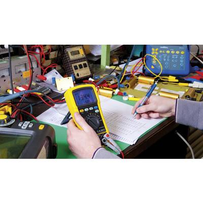 Een persoon meet spanning met een multimeter aan een elektronisch apparaat terwijl hij/zij aantekeningen maakt. Tafel vol kabels en apparatuur.