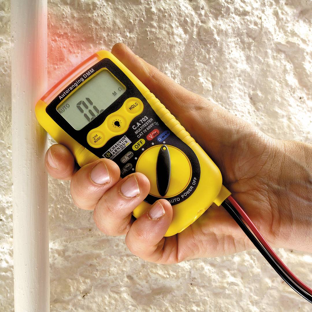 Een hand houdt een geel digitaal multimeter vast dat een spanning van 10,0 mV meet aan een witte buis.