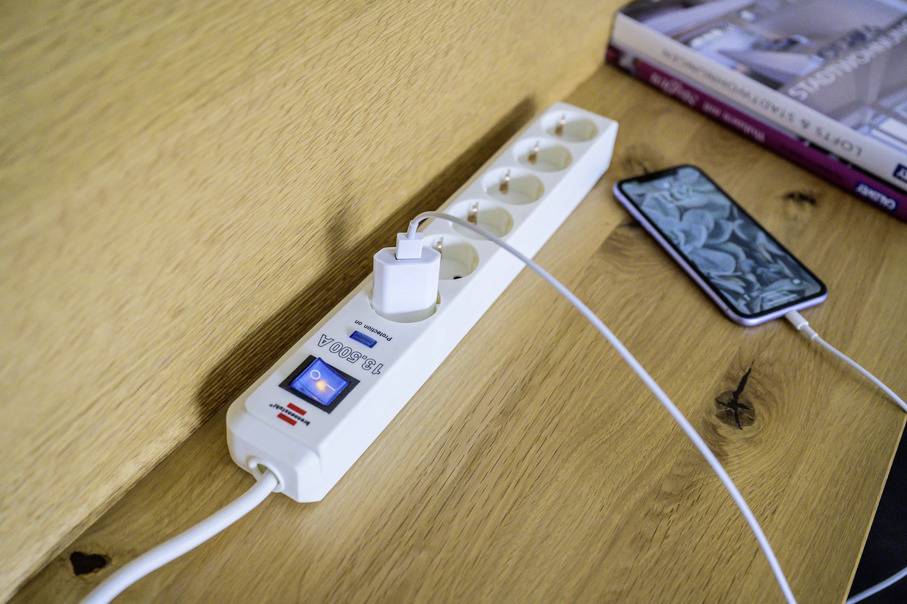 Een witte stekkerdoos met vier vrije en twee bezette stopcontacten ligt op een houten tafel. Een smartphone wordt opgeladen.
