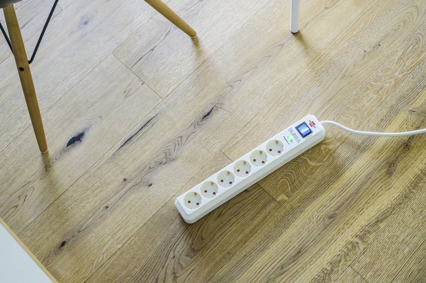 Witte stekkerdoos met vijf aansluitingen ligt op een lichte houten vloer. Een stoelpoot is zichtbaar in de bovenhoek.