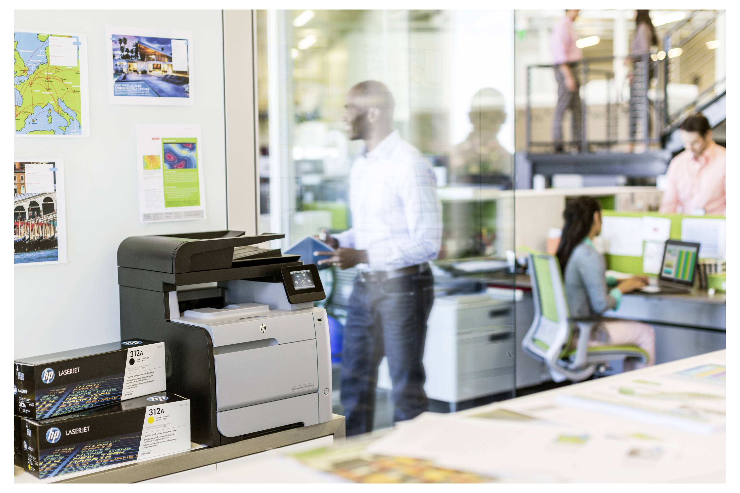 Moderni kantor met een grote printer op de voorgrond en personeel dat aan bureaus achter glazen wanden werkt