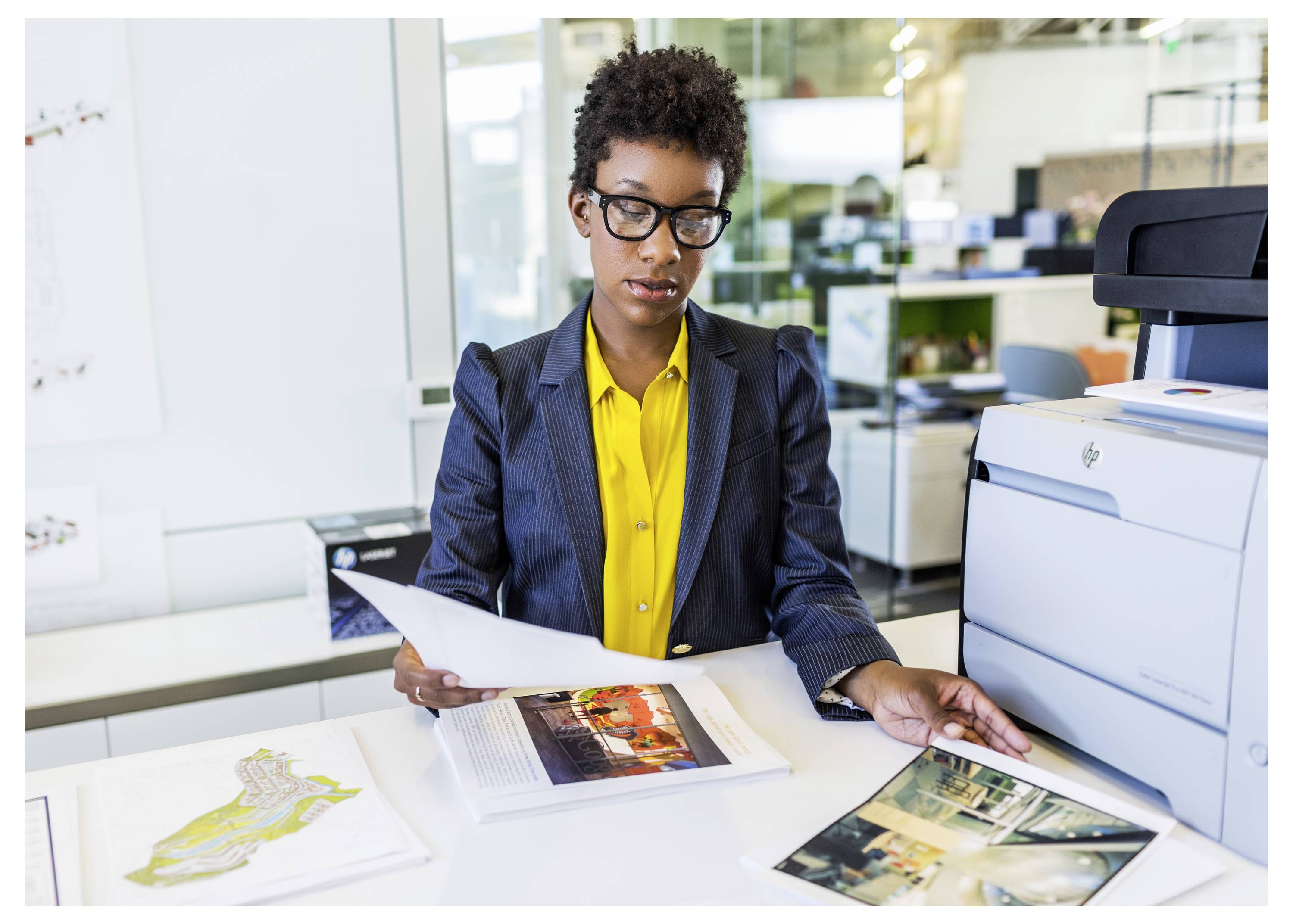 Vrouw in een kantoor bekijkt gedrukte documenten naast een grote printer, terwijl ze pagina's op een bureau onderzoekt met ontwerpproeven uitgespreid.